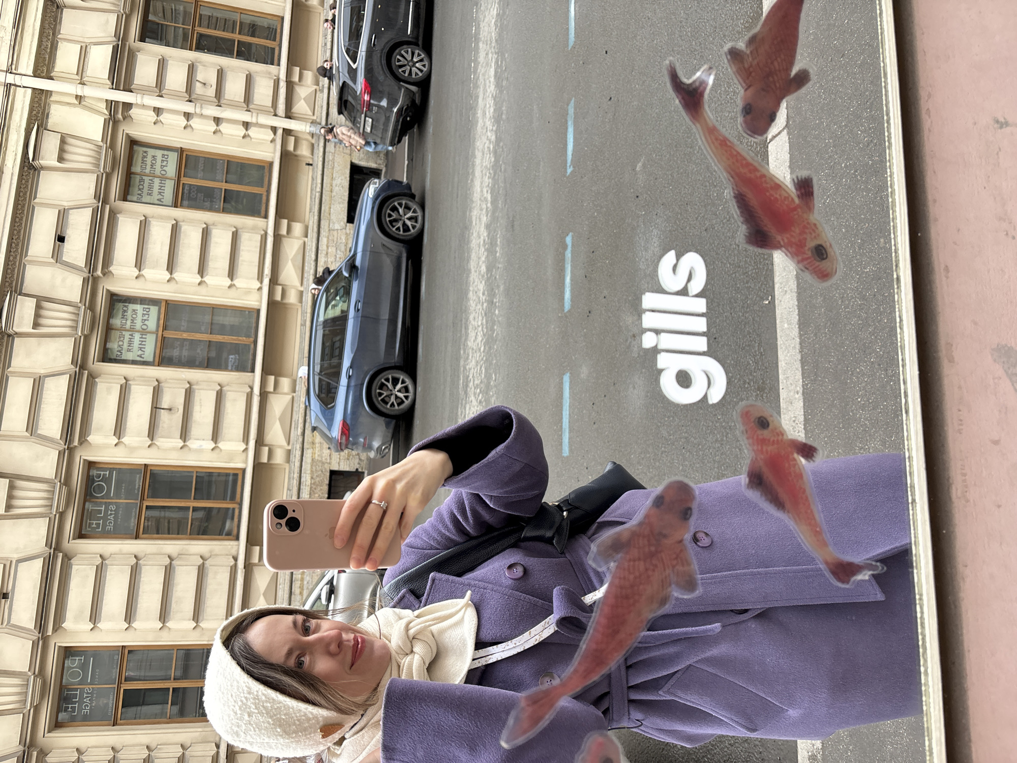 A woman wearing a white head covering and a purple coat takes a selfie in front of a mirror. The mirror reflects the word "gills" on the ground, along with cartoon fish images. Cars and a building with many windows line the street in the background. The building has text in Russian on the windows. Based on the architecture and the Russian text, the location is likely Saint Petersburg, Russia.