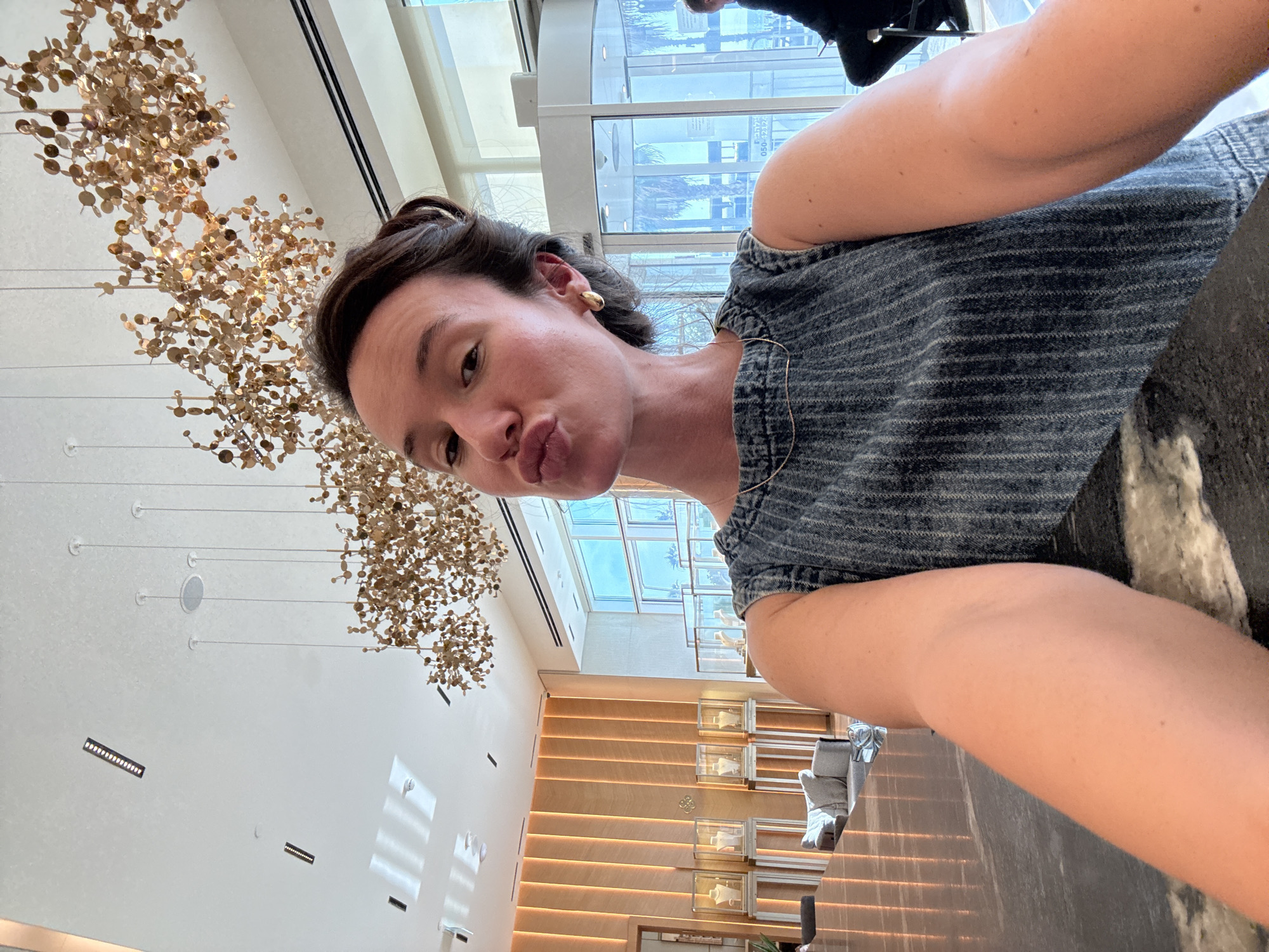 In a well-lit interior, a woman with dark hair pulled back takes a selfie. She is wearing a sleeveless denim top with vertical pinstripes and a thin gold necklace. Her mouth is pursed, and she has a gold earring in her visible ear.
The background suggests a high-end establishment, possibly a jewelry store or a hotel lobby, located in Tel-Aviv, Israel. A large, modern art light fixture with gold, disc-like elements hangs from the white ceiling. To the left, a wall is decorated with vertical wooden panels, illuminated from behind. Several glass display cases, each featuring jewelry on stands, are visible against this wall. In the middle ground, a grey sofa with a textured pillow rests on the floor.
The foreground is dominated by a dark, reflective countertop. Behind the woman, a revolving glass door is visible, with palm trees and blue skies seen through it, indicating an outdoor view. A person in a black outfit is seated to the right, partially visible. The overall impression is one of luxury and modern design.