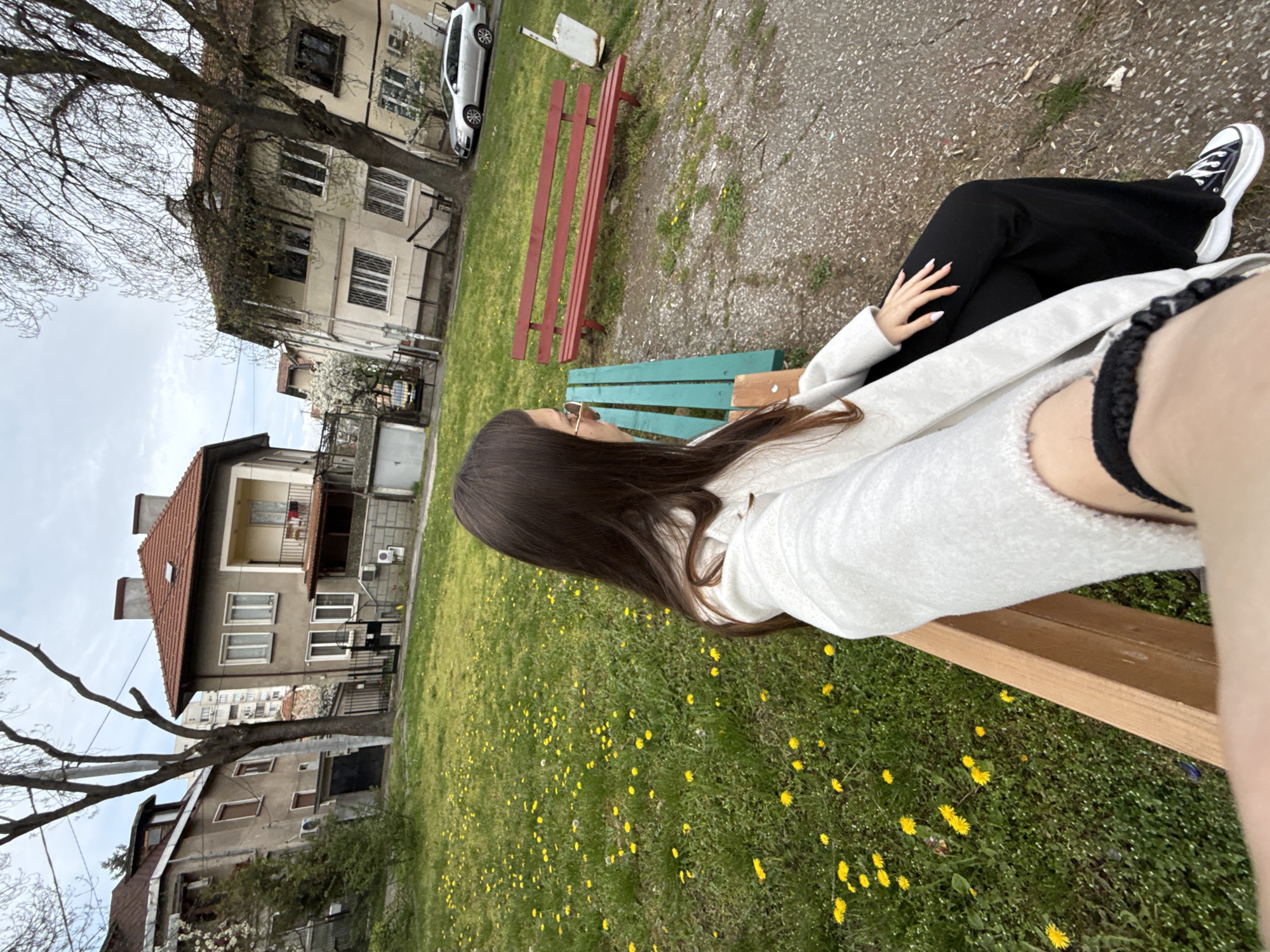 A young woman with long, dark hair is seated on a teal-colored wooden park bench, viewed from a slightly elevated, side perspective. She wears a light-colored (likely white or off-white) long-sleeved top, black trousers, and black and white high-top sneakers. Her left hand, adorned with long, light-colored nails, rests on her leg. She is looking towards the left side of the frame, and a pair of glasses is visible on her face.
The setting is an urban park or green space, with a vibrant green lawn dotted with numerous yellow dandelions. In the mid-ground, another, older-looking red wooden bench is visible. Behind the benches, there's a paved area and several residential buildings, characteristic of a city like Sofia, Bulgaria. These buildings vary in style, some with multiple stories and balconies, and one with a distinctive reddish-brown tiled roof. A white car is parked near one of the buildings, and a tall, bare-branched tree stands prominently on the left side, indicating early spring or late autumn. The sky is overcast. No visible text is present in the image. The scene captures a quiet, casual moment outdoors.