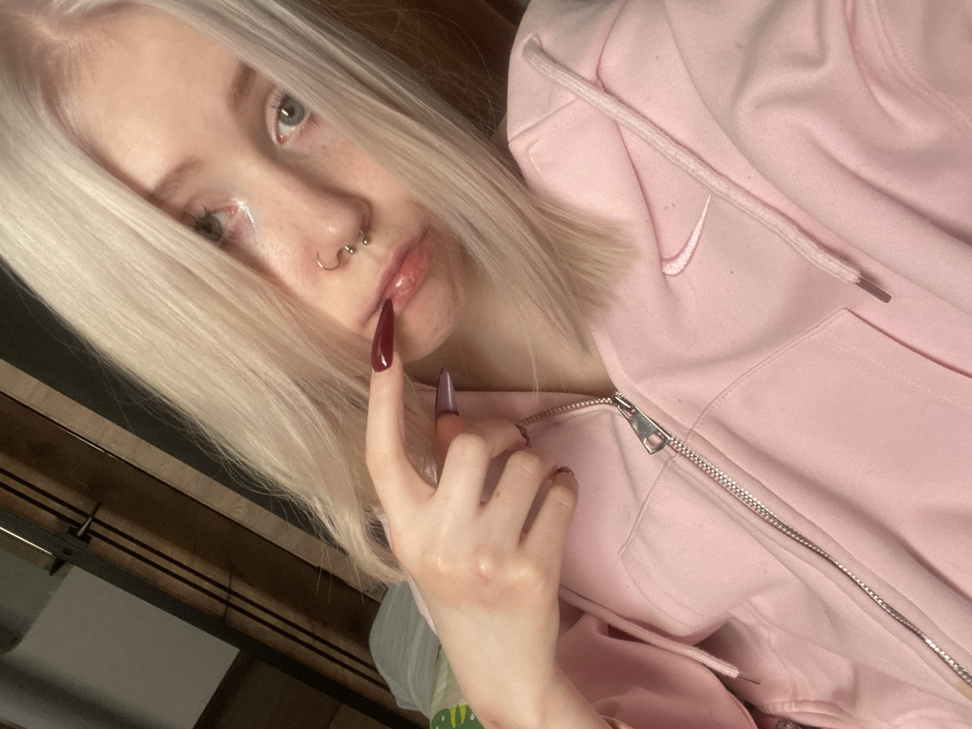 This media file, originating from Izhevsk, Russia, presents a close-up indoor portrait of a young person. The subject has very pale skin, long, straight light blonde hair that frames their face, and striking light-colored eyes, possibly blue or grey. Their gaze is directed towards the camera, conveying a neutral yet somewhat thoughtful or alluring expression.
The person features multiple facial piercings, including a silver stud on one nostril, a silver hoop on the other, and a silver septum piercing. Their lips are slightly parted, with a subtle shine suggesting lip gloss or natural moisture. One hand is positioned near their face, with a single, long, dark red or burgundy-painted fingernail gently touching their bottom lip.
They are wearing a light pink zipped hoodie. A white embroidered Nike swoosh logo is visible on the left side of the chest, indicating the brand. The metal zipper and a matching pink drawstring from the hood are also clearly seen.
The background is indistinct but appears to be an indoor setting, with blurred wooden elements that suggest furniture, such as a headboard or shelving. The lighting is soft and even, indicative of an indoor environment without harsh shadows. No specific time of day or external weather conditions are discernible from the image. The overall impression is a casual, personal shot focusing on the individual's distinctive style and features.