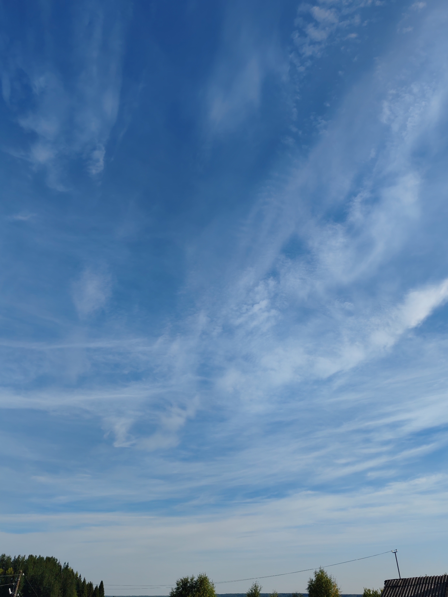 A wide-angle, eye-level shot captures a vast expanse of blue sky with thin, wispy clouds stretching horizontally across the frame. The sky occupies the majority of the image. In the lower portion of the photograph, a dark green line of trees runs from the left edge towards the center. To the right of the trees, a low-lying structure with a dark, corrugated roof is visible. A thin black wire stretches diagonally from the left side of the image across the bottom, supported by poles and disappearing behind the roof of the structure. The overall impression is of a rural landscape under a bright, partly cloudy sky. The location is identified as Chukhlem, Russia.