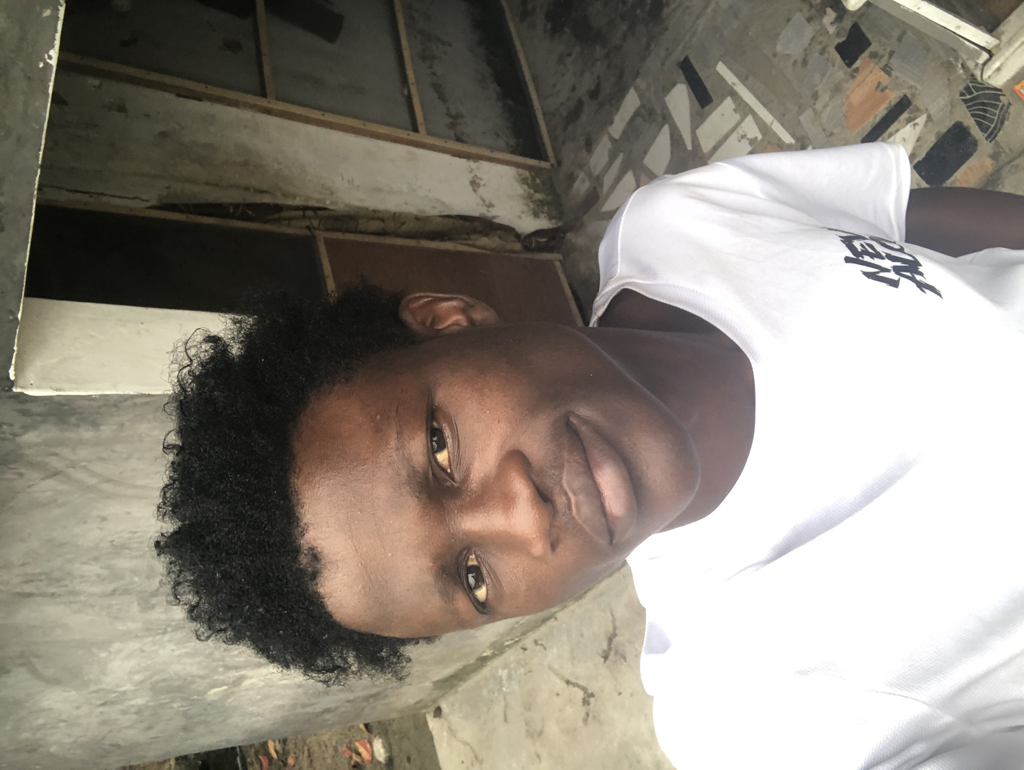 In Ado, Nigeria, a young man with dark, curly hair and brown skin poses for a selfie outdoors. He is wearing a white t-shirt with a black graphic design on the left side of his chest. The background consists of a rough, textured gray wall on the left, and on the right, a partially open doorway revealing a dark interior with exposed wooden beams and a corrugated metal roof. The ground is paved with irregular geometric shapes in shades of gray, white, and black. To the left of the man, near the gray wall, a few fallen brown leaves are visible on the ground. The lighting suggests it is daytime, with some shadows indicating overhead light.