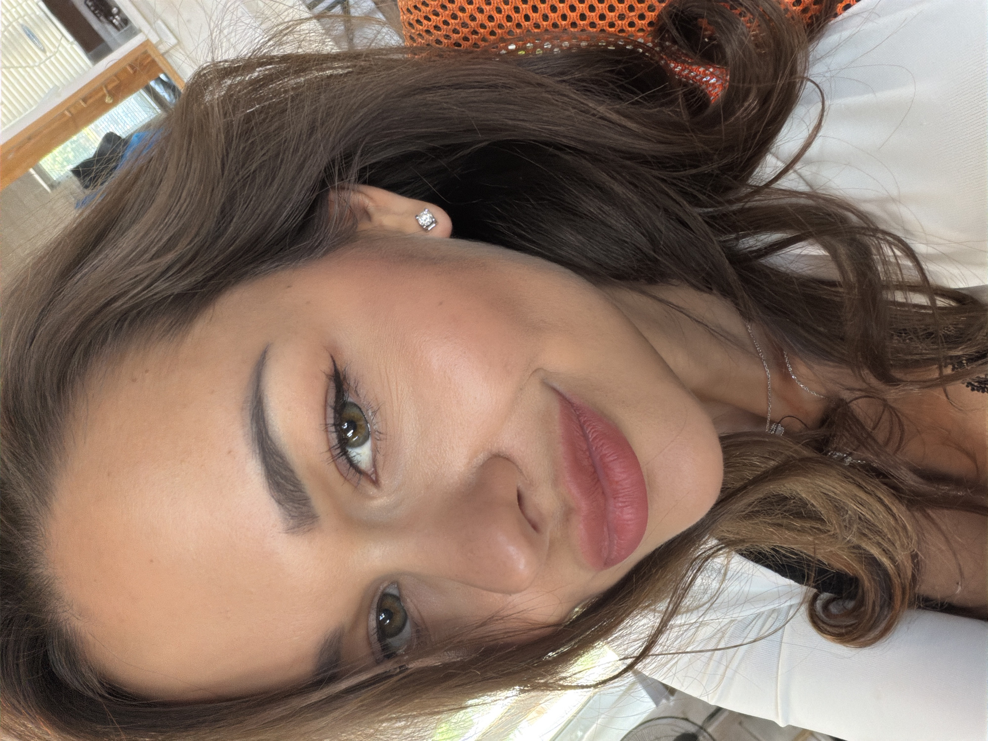 A close-up selfie shows a woman's face and upper chest. She has long, wavy brown hair that frames her face and falls over her shoulders. Her eyes are green, and she is wearing eye makeup, including eyeliner and mascara. She has full lips with pink lipstick. She is wearing a white top and a silver necklace. A diamond stud earring is visible in her right ear. The background is blurred, but appears to be an indoor setting with a window and blinds. No location in Saint Petersburg, Russia can be determined from the image.