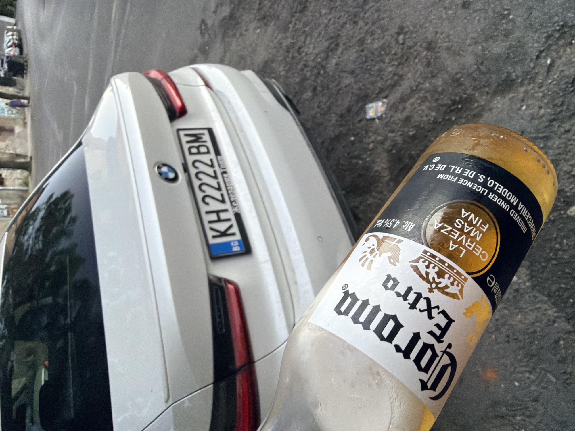 The image shows the rear of a white BMW sedan parked on an asphalt street in Dupnitsa, Bulgaria. The license plate reads "KH 2222 BM" with a blue and yellow BG sticker on the left. In the foreground, a partially visible bottle of Corona Extra beer is held at an angle, obscuring part of the car. The street is lined with trees and buildings in the background, and a person in a purple shirt is visible walking away from the camera.