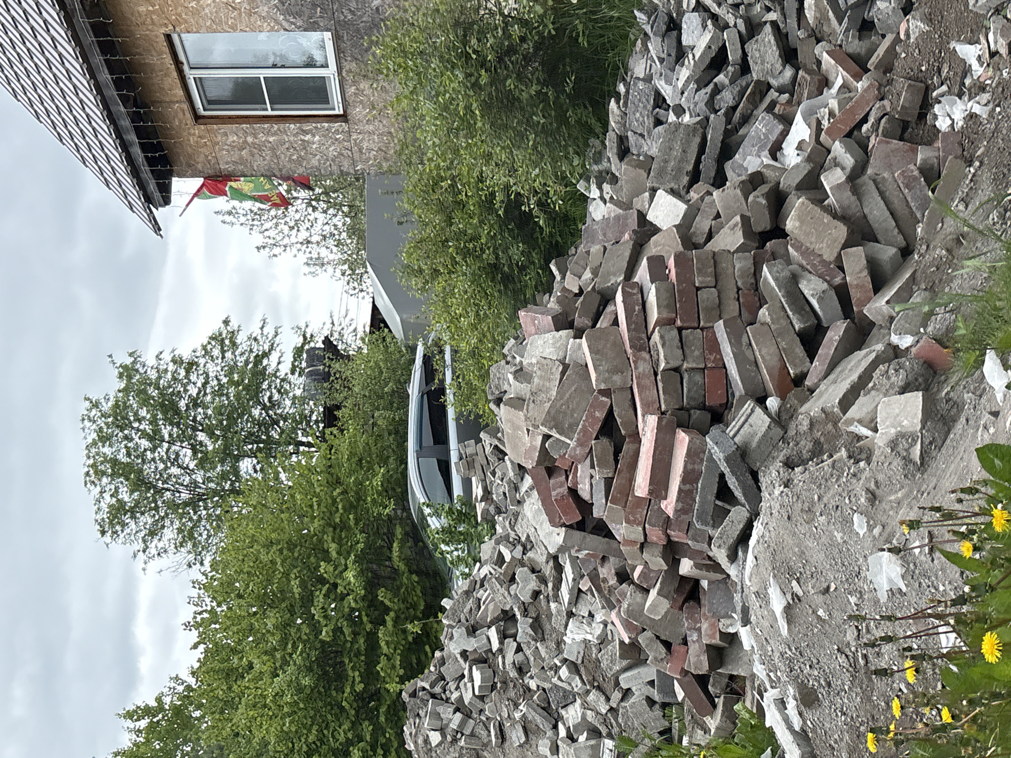 The image depicts a pile of mixed bricks and concrete debris in Озон Снт, Russia. The pile is predominantly gray and red, consisting of whole and broken bricks, as well as fragments of concrete. Vegetation, including green bushes and yellow dandelions, is visible nearby. In the background, there's a partial view of a house with a light-colored, possibly wooden, exterior and a window. A vehicle, likely a car, is partially visible behind the debris. The sky appears overcast. A flag is visible, hung outside the house.