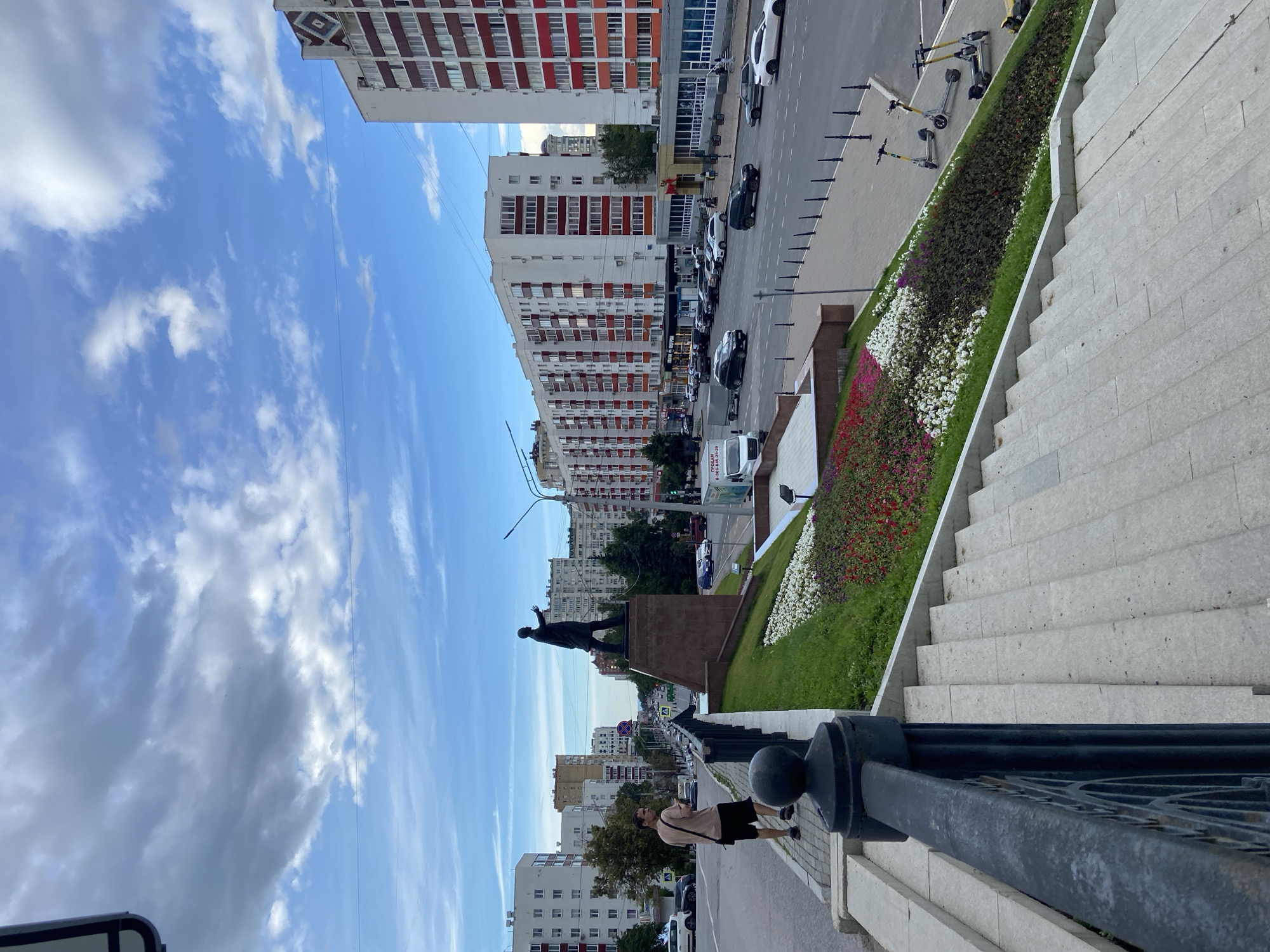 A medium-shot, eye-level, outdoor photograph captures a street scene in Ufa, Russia. In the foreground, a person stands on a sidewalk, facing left, looking towards a statue of Lenin. The statue, a prominent dark bronze figure, stands on a large stone pedestal. Behind the statue, a multi-lane road carries traffic. The buildings lining the street are modern, tall, and brightly colored, with white and red facades. Sidewalks, streetlights, and a flower bed with a variety of colored flowers are also visible. The sky is blue with scattered clouds, indicating a daytime scene. Two electric scooters are parked on the sidewalk next to the flower bed.