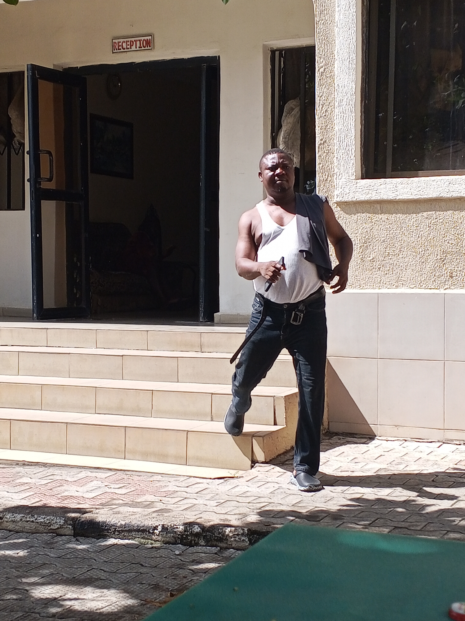 An adult male with dark skin is captured in motion, stepping down or having just stepped off the tiled stairs in front of a building. He is wearing a white ribbed tank top, dark jeans with a dark belt and silver buckle, and dark athletic shoes. A dark piece of clothing or fabric is draped over his left shoulder, and he holds a slender, dark object in his right hand. His expression is neutral as he looks towards the camera.
The setting is outdoors, in bright daylight, indicated by strong shadows cast on the ground and building. Behind the man is a light-colored building with a textured facade. A prominent, open double doorway with dark frames leads into the building. Above this entrance, a red sign clearly reads "RECEPTION." Tiled steps lead up to this doorway. To the right of the entrance, a large window with dark frames is visible. The interior seen through the open door is dimly lit, revealing a framed picture on a wall and some dark furniture.
The ground in the foreground features patterned paving stones, a concrete curb, and a different type of patterned brickwork. A green surface, likely a table, is partially visible in the bottom right corner. The overall scene suggests the entrance to a public building, possibly a hotel or guesthouse, in a sunny environment.