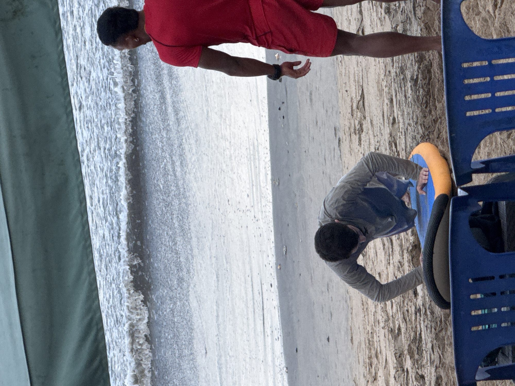 Two individuals are present on a sandy beach in Kuta, Indonesia. The person in the foreground, dressed in a gray long-sleeved shirt and dark pants, is in a push-up position with their hands on a blue and yellow surf mat. They are facing away from the camera, towards the ocean. To the right, a person wearing a red t-shirt and red shorts stands facing the ocean, observing the other individual. The ocean waves are visible in the background. Two blue plastic chairs are positioned in the lower foreground, partially obscuring the sand. A green awning stretches across the top of the image.