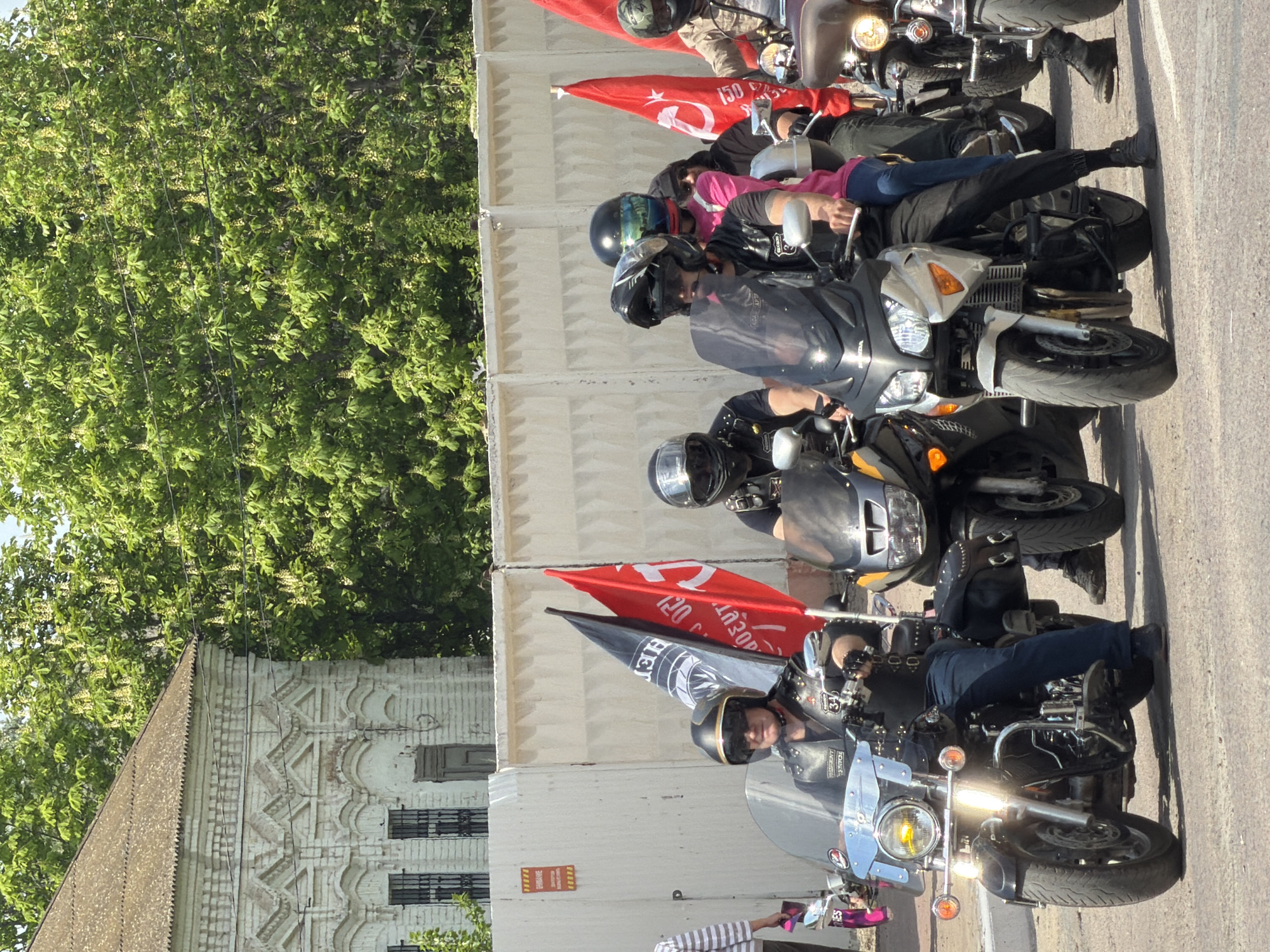 The image shows a group of motorcyclists gathered on a street in Kamyshin, Russia. The riders are astride their motorcycles, which are parked in a row along the side of the road. The riders are wearing motorcycle helmets and protective gear. Some of the motorcycles have flags attached to them. The environment includes an ornate white building with barred windows and a large tree full of leaves.