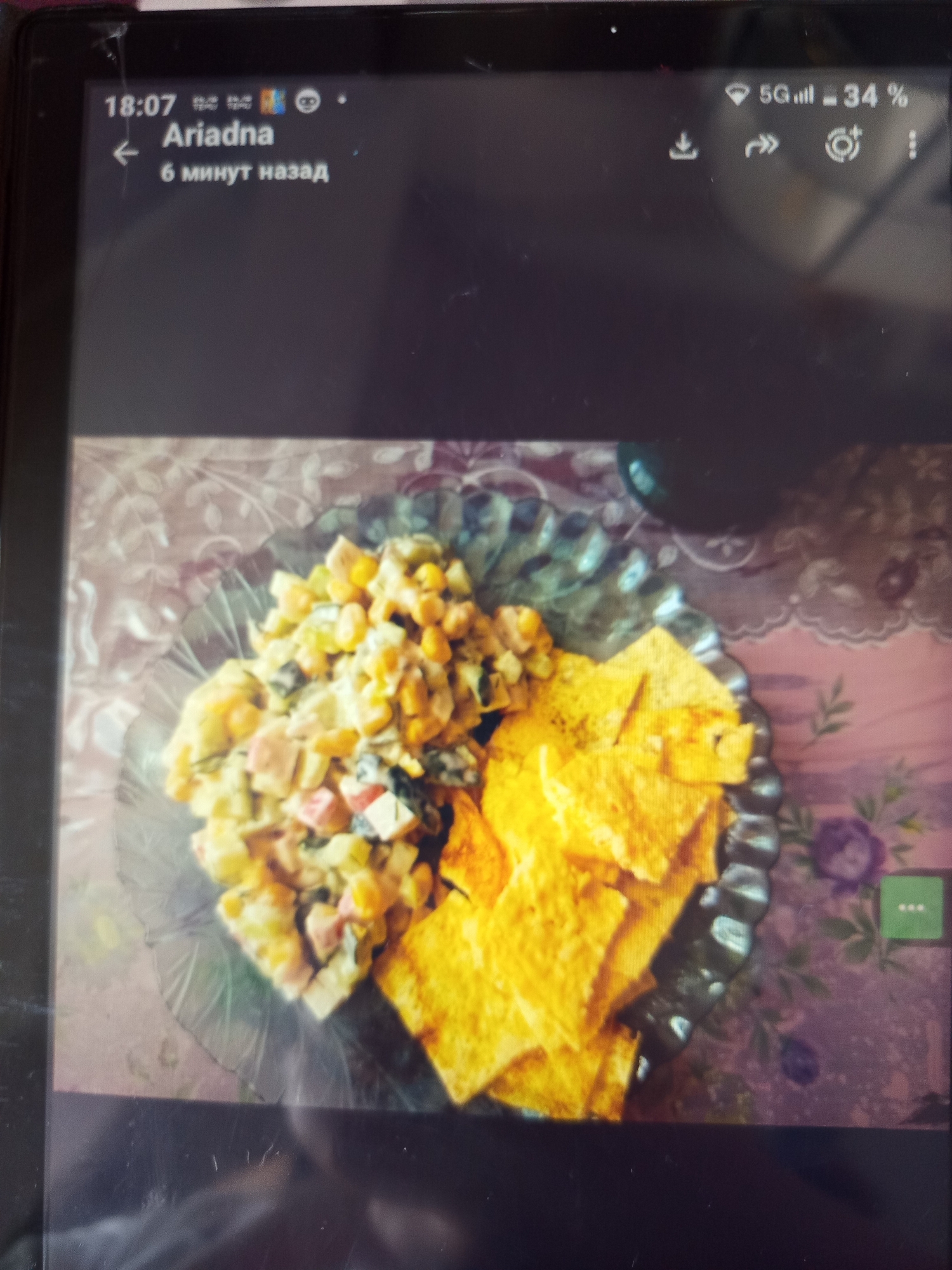 The image displays a close-up of a plate of food, likely taken indoors. The food consists of a salad mixture containing corn, diced vegetables, and possibly meat, arranged on the left side of a clear glass plate. To the right of the salad are several pieces of what appear to be tortilla chips or some other type of fried flatbread. The plate is placed on a surface with a patterned tablecloth featuring floral designs in shades of pink and purple. The lighting suggests an indoor environment, possibly a home or restaurant. The upper portion of the image shows a tablet screen with a time stamp "18:07" and the name "Ariadna" with text below indicating "6 минут назад" (6 minutes ago). Additionally, a status bar shows "5G, signal strength, and 34% battery". There are icons for download, reply, and settings. The context suggests this is a captured image from a device, possibly a screenshot or a photo taken of a screen. There is no indication of the specific location within Bishkek City, Kyrgyzstan.