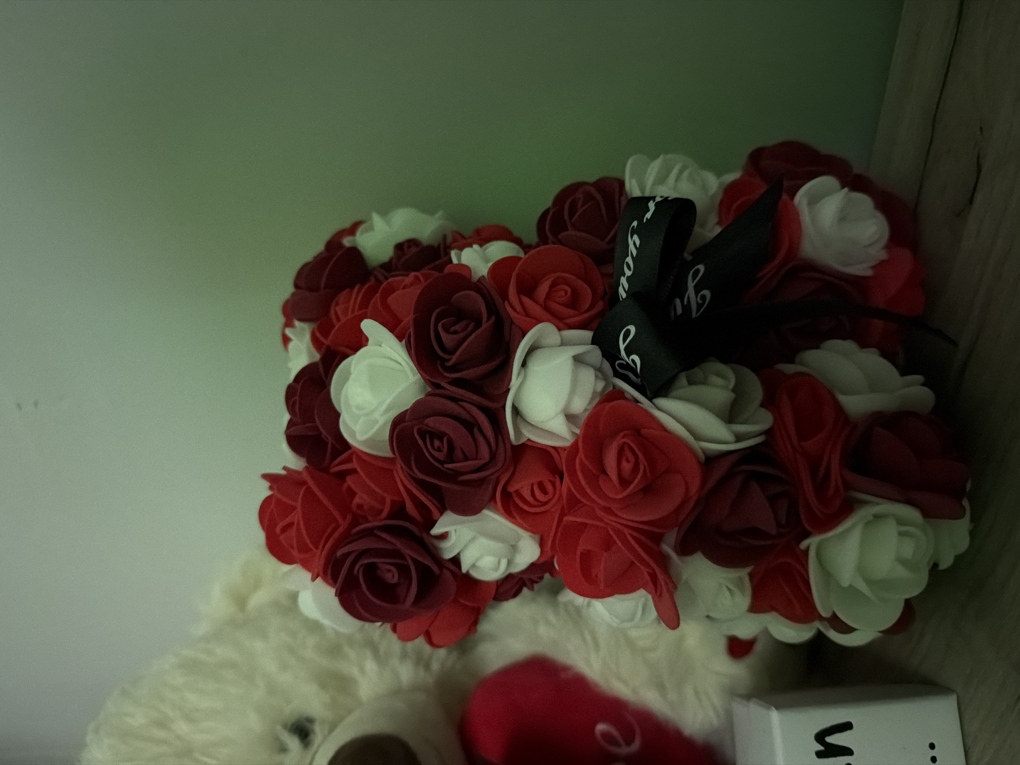 The image displays a static indoor arrangement on a wooden surface positioned against a plain, light greenish-grey wall. The primary subject is a teddy bear-shaped floral arrangement, meticulously constructed from numerous artificial roses in shades of red, dark red, and white. A black ribbon with partially visible white text is tied around the arrangement's neck. To the left, the upper portion of a white, fluffy teddy bear is visible, holding a red heart-shaped object that displays a partial white letter 'e'. Adjacent to the white teddy bear, a white rectangular block or box is present, showing partial black text 'n...' on its visible side. The wooden surface exhibits horizontal plank lines and grain patterns. No individuals are present, and no actions or interactions are depicted. The specific location within Varna, Bulgaria, is not discernible from the visual information provided.