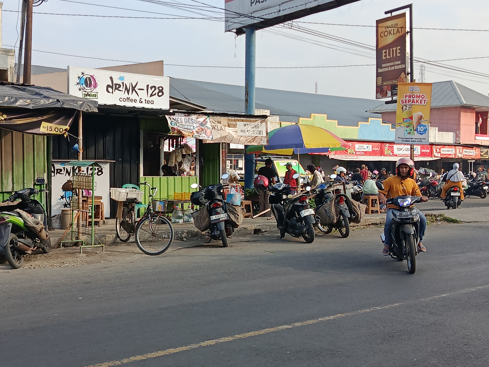 The image shows a street scene in Kalijeruk, Indonesia, with a focus on a row of shops and stalls. In the foreground, a man on a dark scooter, wearing an orange shirt and blue jeans, rides down the asphalt road, which has a yellow lane marker. Behind him, several other scooters and a bicycle are parked along the roadside, some laden with goods.
The shops are varied in appearance. One prominent stall has a sign that reads "DRINK-128 coffee & tea." Next to it, a vendor stall is visible with items displayed for sale, including bags and possibly food. Further down the street, colorful awnings and umbrellas are set up over other stalls.
A tall signpost in the background displays advertisements, including one for "COKLAT ELITE" and another for a "PROMO SEPAKET." The architecture of the buildings behind the stalls suggests a commercial or market area. The overall atmosphere appears to be a busy, everyday street scene in a commercial district. The lighting indicates it is daytime, with sunlight casting shadows.