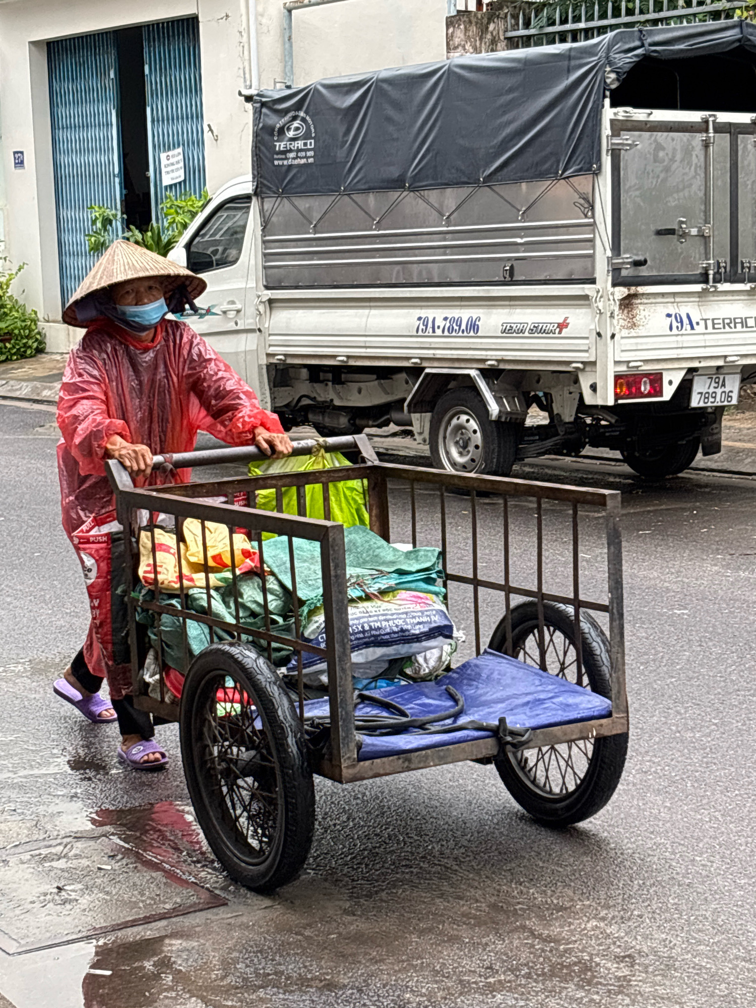 A person wearing a red raincoat, a conical hat, and a face mask pushes a metal cart filled with various items, including colorful bags and green tarps. The cart has two large spoked wheels. The person is walking on a wet asphalt street, indicating it is raining or has recently rained. In the background, a white truck with a black tarp covering its cargo area is parked or moving slowly. The truck has license plates and text printed on its side. To the left of the person is a building with blue metal doors and some greenery. The scene appears to be a street in Nha Trang, Vietnam, suggested by the style of the vehicle, the conical hat worn by the person, and the presence of a building with characteristic architecture.