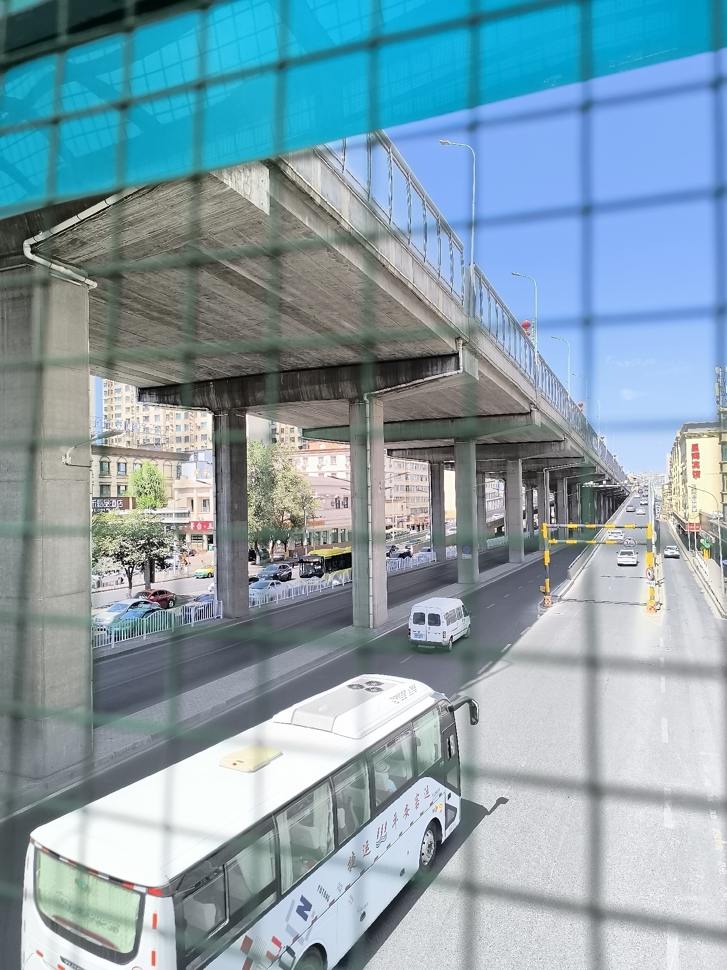 The image depicts an elevated roadway passing over a street in Ürümqi, China. The foreground is partially obscured by a green metal mesh. A white bus is traveling on the lower street, with a white van behind it. The elevated roadway is supported by concrete pillars and has a bright blue roof structure partially visible at the top. The street below is lined with buildings and trees. Cars are also visible on the street and on the elevated roadway. The sky is clear and blue. The scene appears to be a busy urban environment.