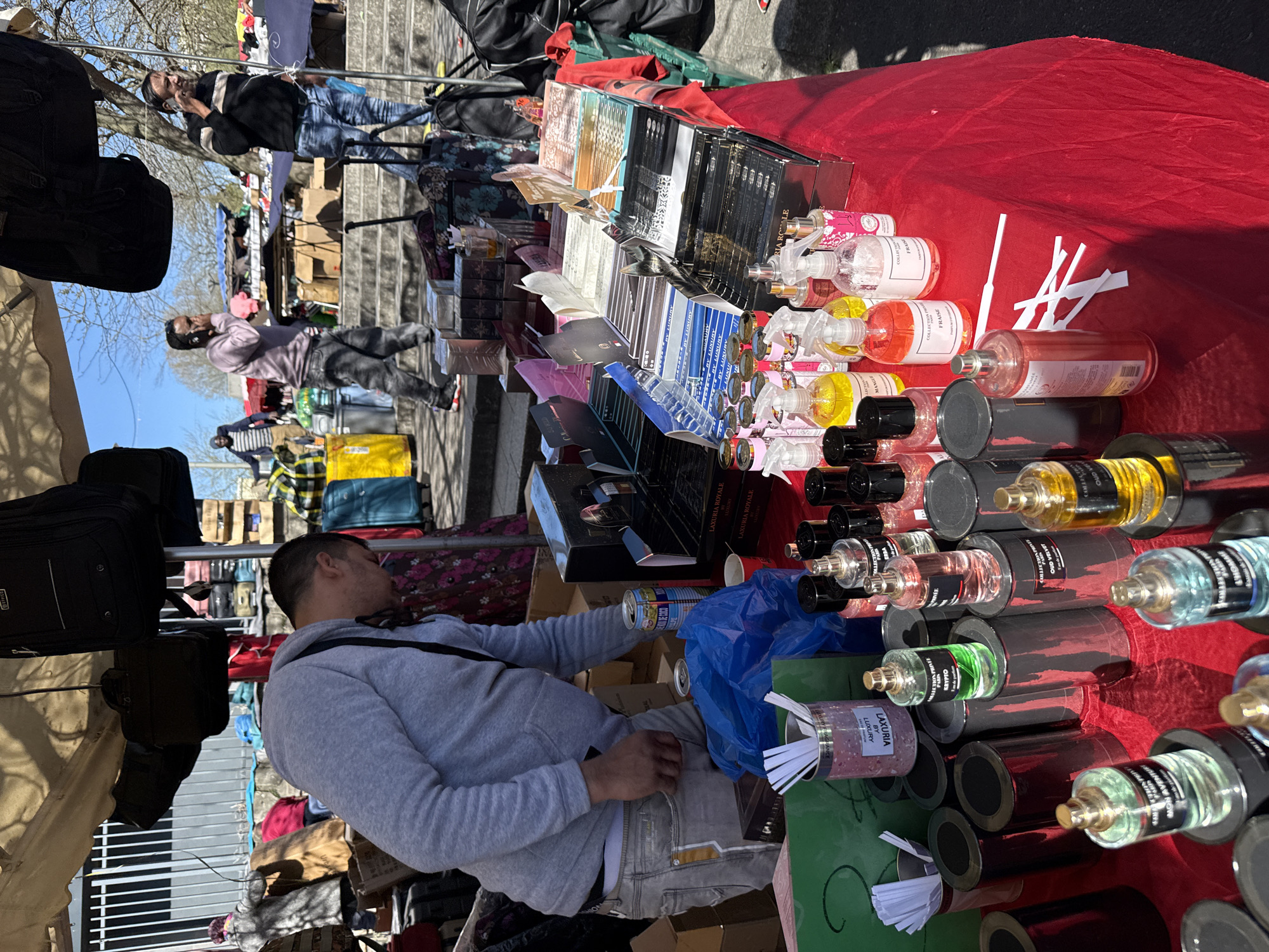 The image captures a scene at an outdoor market. A vendor, dressed in a gray hoodie and jeans, sits behind a table draped with red fabric, which displays an assortment of perfumes and fragrances in various bottles and packaging. To the vendor's left, a row of black bags hangs from a rack. In the background, people stroll along a walkway beside a flight of stairs, suggesting a moderately bustling environment. The products on the table feature labels like "LAXURIA," "OUD VELVET," "FRAISE," and "COLLECTION PRI." Based on the visible clues, the market appears to be located in Garges-lès-Gonesse, France.