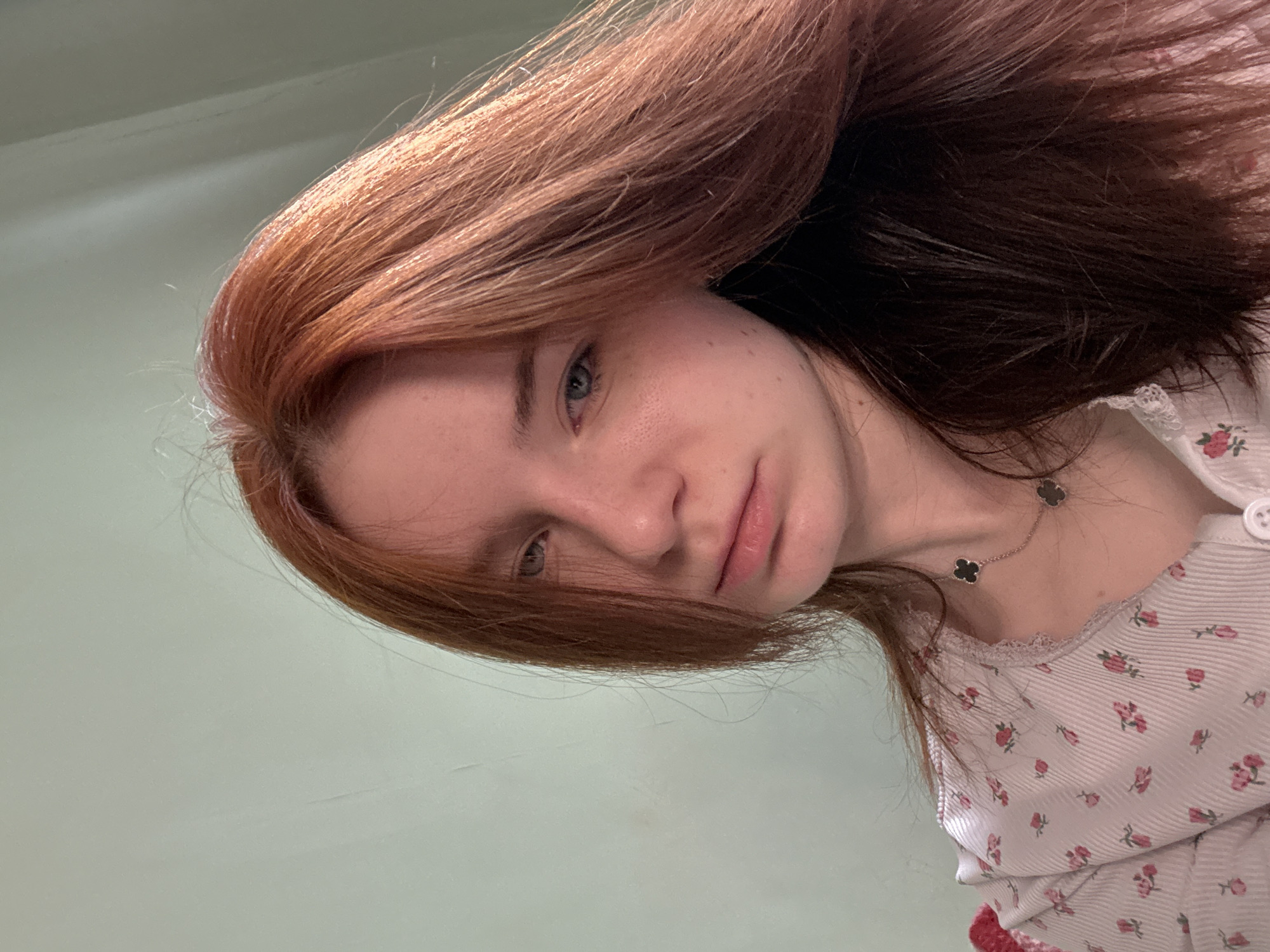 A close-up, angled selfie photograph of a young woman with red and dark brown hair. She has fair skin, light blue eyes, and is wearing a light-colored top with a floral print and lace trim around the collar. She also has a necklace with clover-shaped pendants. The background is a plain, light green wall. The location is unknown.