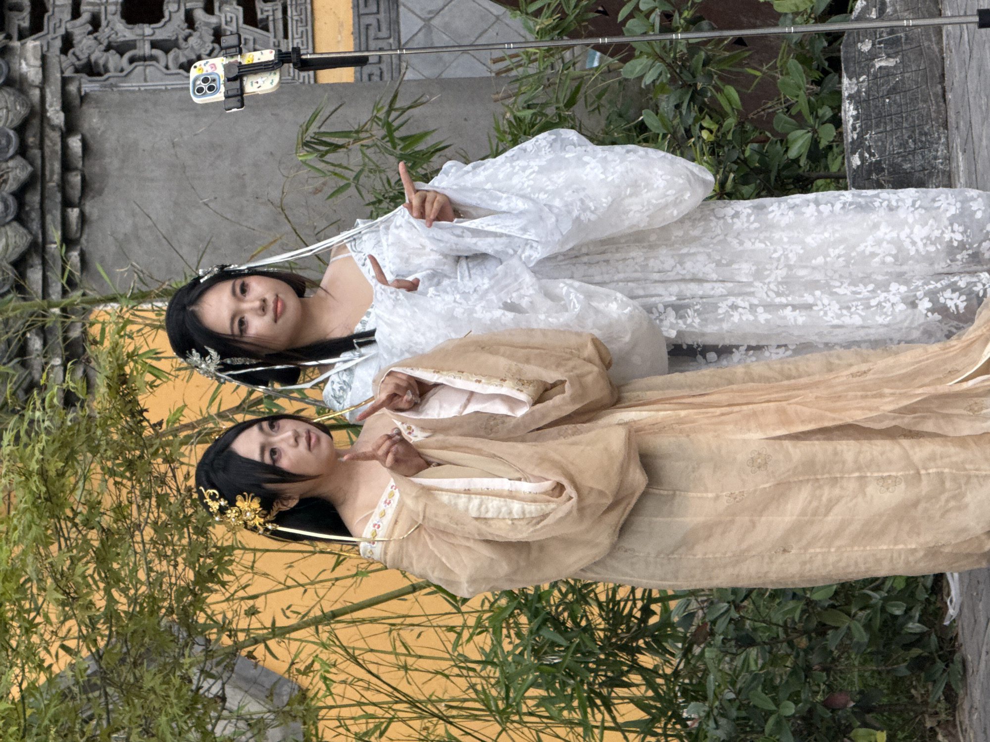 Two Asian women in traditional attire pose for a photo using a smartphone attached to a selfie stick. One woman is dressed in a white floral gown and the other in a beige gown with gold accents. They stand in front of bamboo plants and a wall with traditional Chinese architectural details, possibly in the Yuzhong District of China. Both women are making hand gestures.