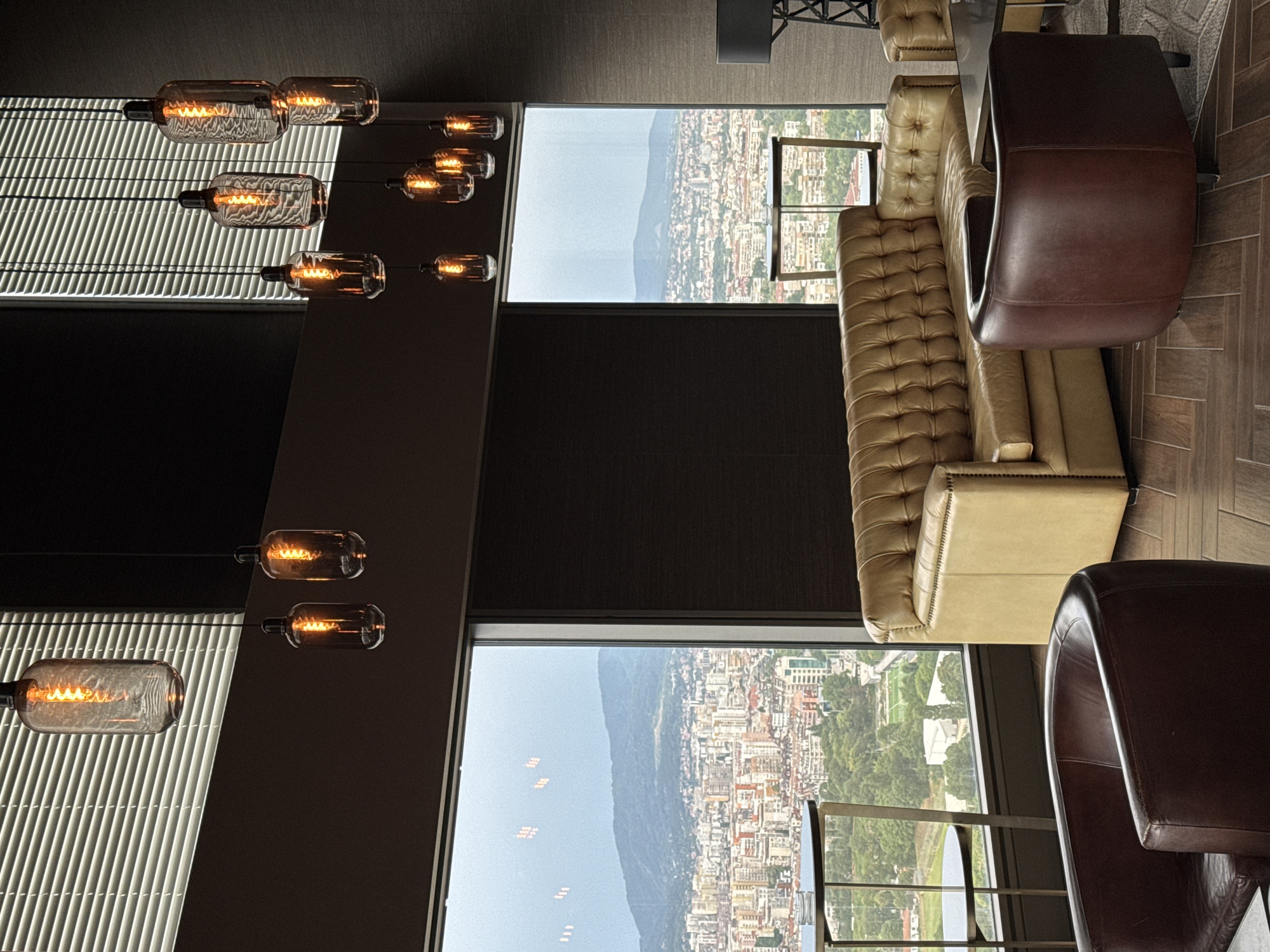 This image depicts a sophisticated interior with a panoramic view of Sofia, Bulgaria. The foreground features luxurious leather furniture: a deep brown armchair with a curved back and a tufted camel-colored sofa. The sofa is positioned to overlook a cityscape, with a dark brown, curved armchair placed in front of it. In the background, large windows offer an expansive vista of the city, characterized by a dense collection of buildings with red-tiled roofs, interspersed with greenery. Beyond the city, a range of rolling, verdant hills forms the horizon under a hazy, overcast sky.
The interior design is characterized by modern, industrial-style pendant lights with exposed Edison bulbs, suspended from the ceiling. These lights cast a warm glow. The walls are paneled with dark wood, creating a rich and intimate atmosphere. A small, round table with a metal frame and glass top is visible to the left of the sofa, near the window, with an ashtray on its surface. The flooring is a herringbone pattern of dark wood planks. To the far right, a glimpse of a modern floor lamp with a black shade suggests further contemporary design elements. The overall impression is one of an upscale lounge or restaurant with a commanding view of Sofia.