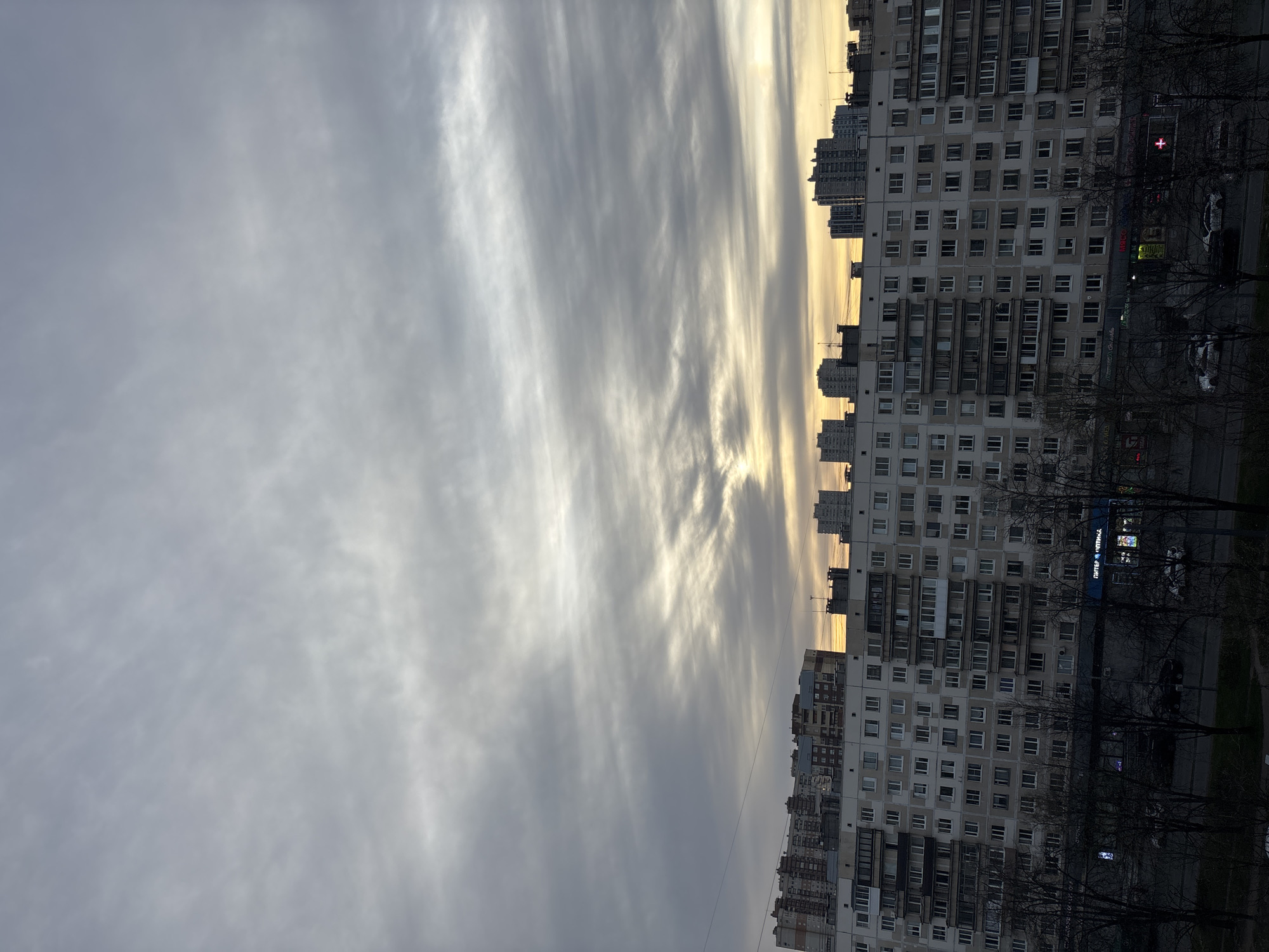 The image shows a cityscape in Unknown, Russia, under an overcast sky. In the foreground, there's a row of tall residential buildings with a repetitive window pattern, suggesting Soviet-era construction. The buildings are densely packed, indicating an urban area. Trees with bare branches are visible in front of the buildings, possibly indicating late autumn or winter. Between the buildings and the sky, there's a golden glow that suggests either sunrise or sunset. The sky is covered with layers of stratus clouds, giving the scene a muted, somber tone. A few parked cars are visible at street level near the buildings. A sign is seen on one of the buildings, but the letters are unreadable. A traffic light with red lights lit up is present.