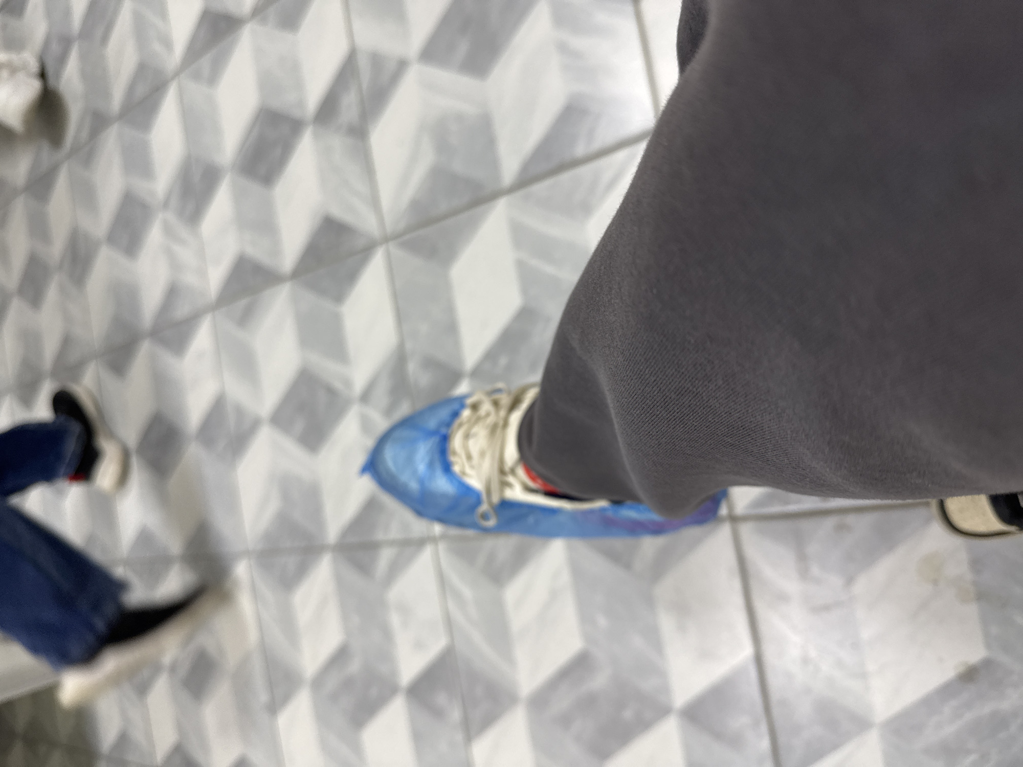 A high-angle shot shows a person's legs and feet on a geometric patterned floor, possibly tile. The person on the right is wearing gray sweatpants and one foot is covered in a blue disposable shoe cover. Another person on the left wears blue jeans and black and white sneakers. A blurred partial view of other individuals and their shoes appears in the periphery of the frame. The location is unspecified, and there is no information to confirm that the image was taken in Anapa, Russia.