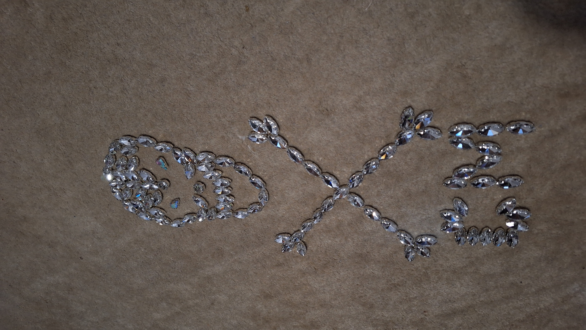 An image displays an arrangement of clear, faceted stones, possibly rhinestones or crystals, spelling out "O X E" on a textured, light brown surface. The background appears to be a soft material, like fabric. The 'O' is formed by an oval shape of stones with a few smaller, rounder stones placed inside. The 'X' is composed of several elongated stones forming a cross shape, and the 'E' is similarly constructed from multiple sparkling stones, presenting a stylized appearance. The stones reflect light, showing subtle iridescent hues. The scene is a static, indoor display, likely photographed under artificial lighting. This arrangement is situated in Blagoevgrad, Bulgaria.
