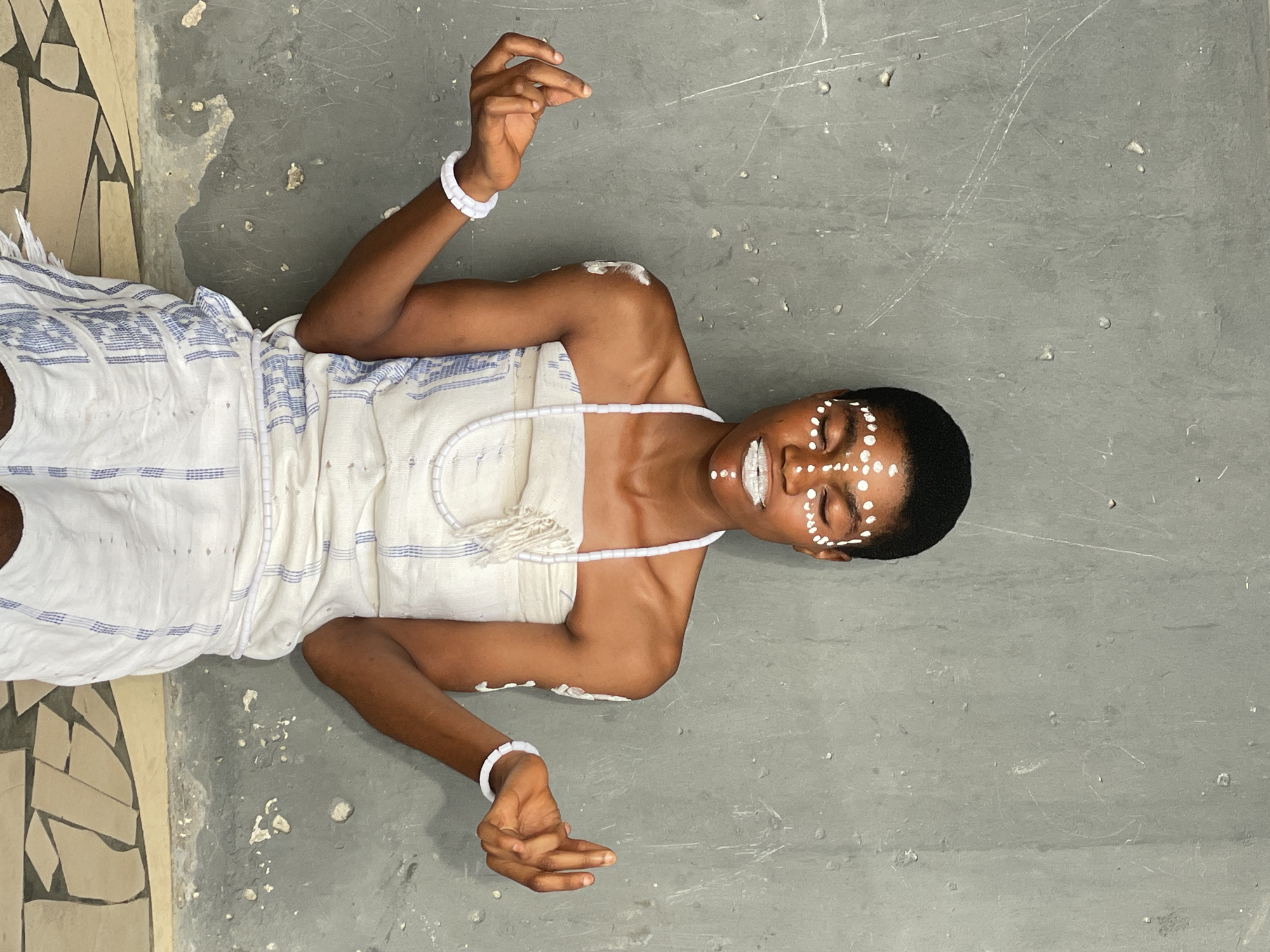 A person is lying on a gray concrete surface, possibly a floor or ground, in what seems to be an indoor or semi-enclosed space. They are wearing a white cloth with blue stripes and white adornments, including a beaded necklace and wrist cuffs. White markings are applied to their face. The setting appears to be a simple, possibly public or unfinished space, part of which includes beige and brown tiles.
While the prompt requests information about Badagry, Nigeria, the image alone doesn't provide specific evidence to confirm this location.