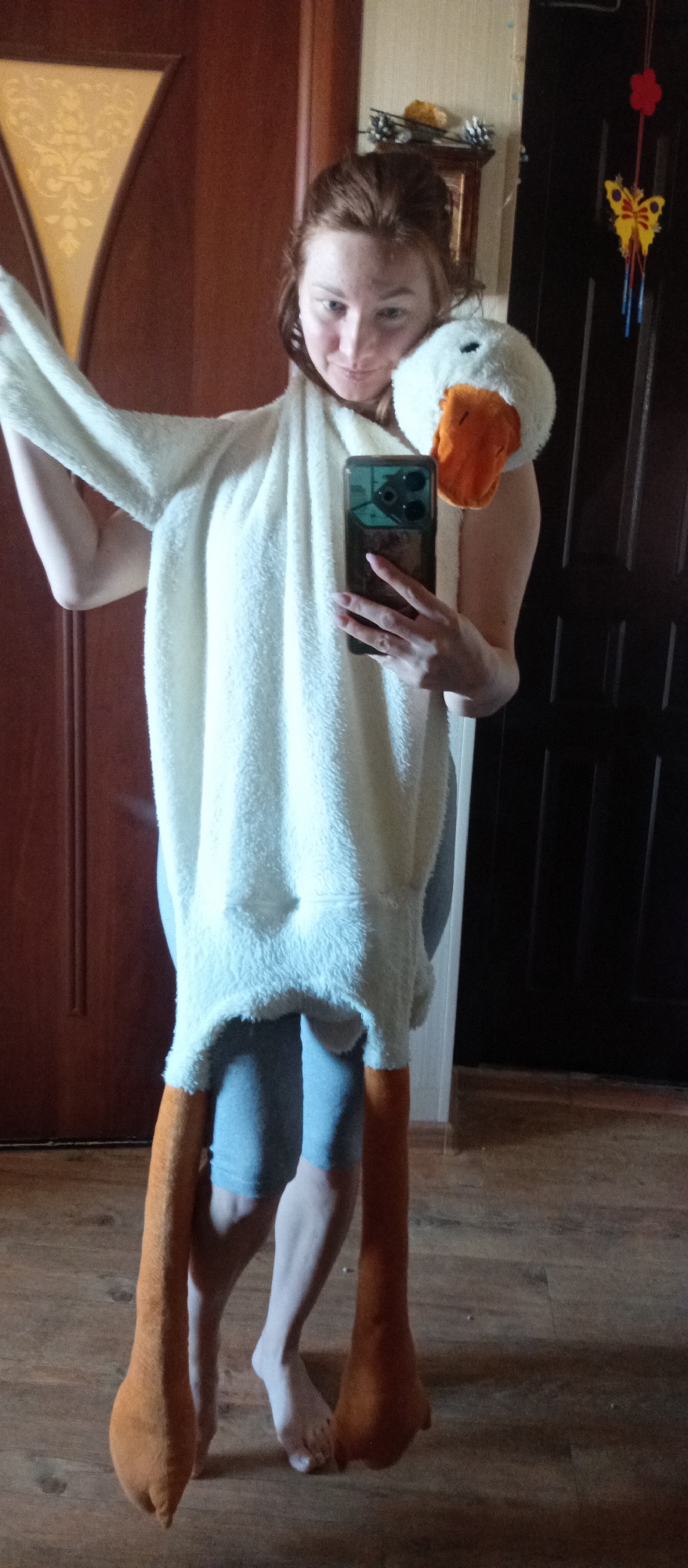 A full-length shot shows a woman standing indoors, holding a cell phone and wearing a white, furry goose costume. The costume has a goose head on her shoulder and long, orange legs that reach the floor. She is wearing gray leggings underneath. She is barefoot. The interior includes two doors - one wooden with a decorative inset, and another dark door with vertical panels. A butterfly decoration is visible on the dark door. The location is Unknown, Unknown.