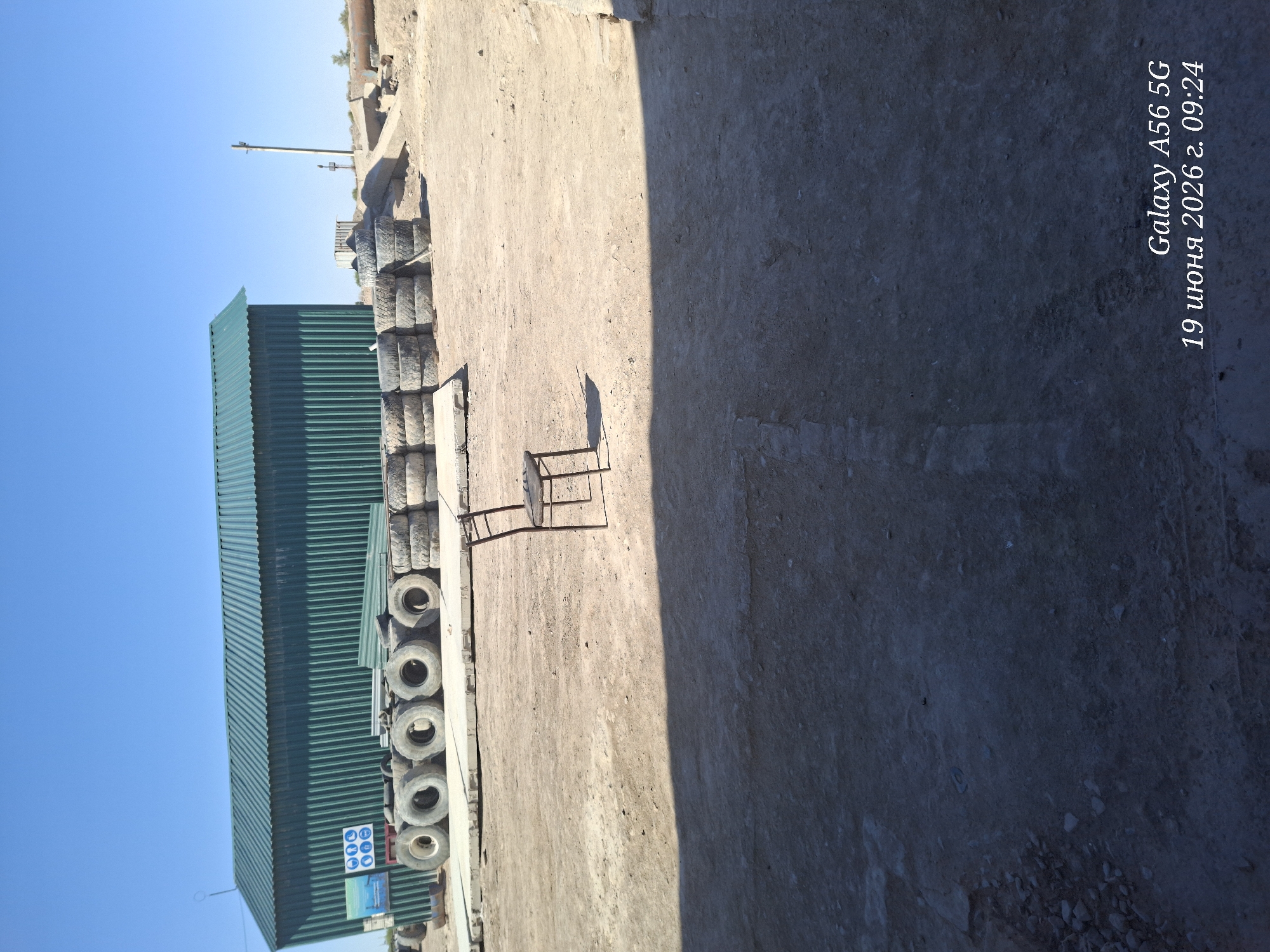 The image depicts an outdoor scene in a dusty, possibly industrial or construction area. A long, green corrugated metal building stands in the background, with a bright blue sky overhead. In front of the building, a row of large tires are stacked or lined up. In the middle ground, a single, empty metal chair with a dark seat is positioned on the dirt ground, casting a shadow. The ground is uneven and appears to be composed of dirt and gravel. In the lower right corner of the image, text reads "Galaxy A56 5G 19 июня 2026 г. 09:24".