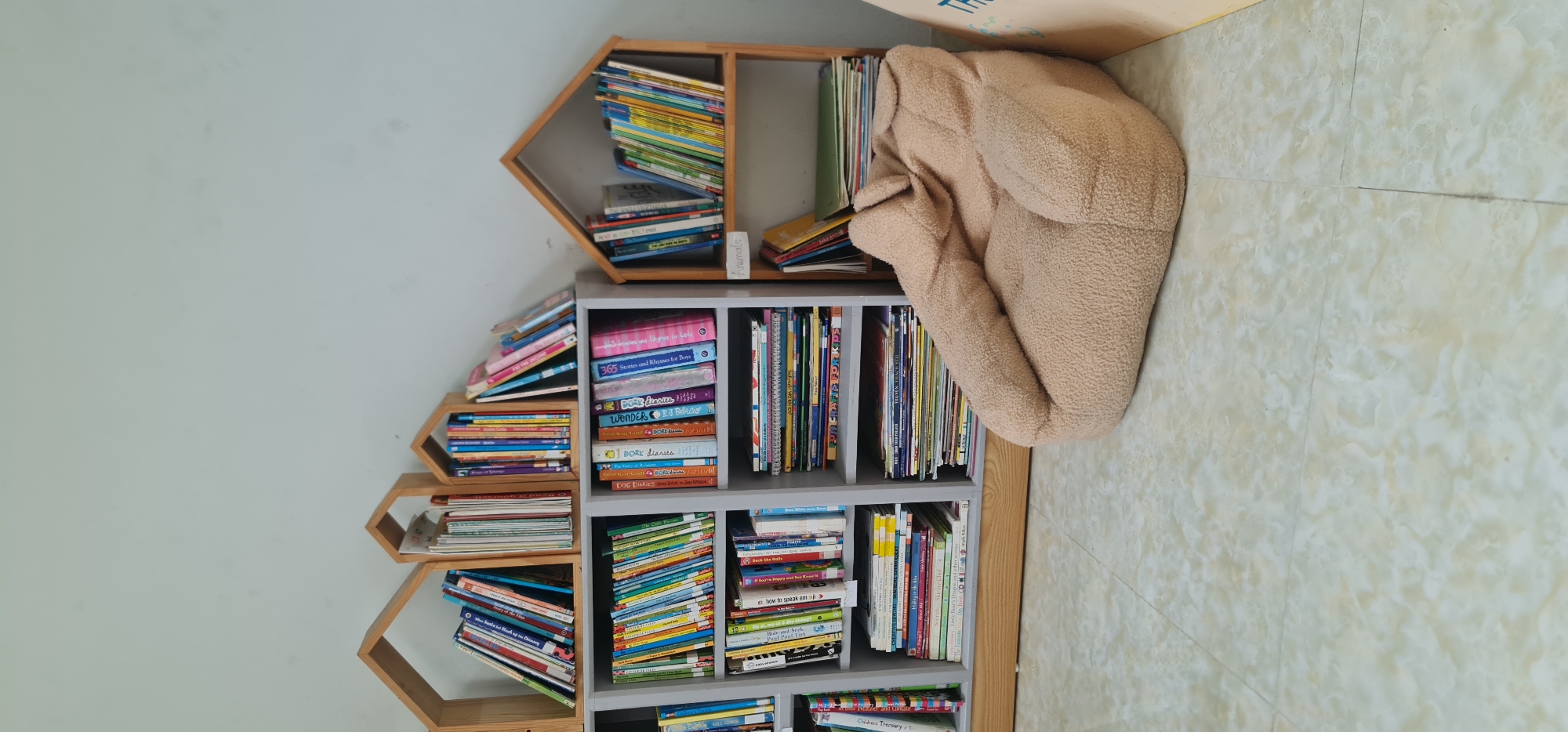 The image shows a bookshelf filled with books, including titles like "365 Stories and Rhymes for Boys," "DORK diaries," "WONDER" by RJ Palacio, "1 DORK diaries," and "DOG DIARIES" by BJ Daniels, as well as "FUN CRAFTS FOR KIDS" and "how to speak emoji". The bookshelf is composed of two differently styled sections: a wooden shelf with house-shaped cubbies and a gray shelf with rectangular cubbies. There is a beige fluffy object, possibly a blanket or a beanbag, next to the bookshelf. The scene is indoors, with a light-colored wall and a tile floor. It is not possible to determine if this image was taken in Đà Nẵng, Vietnam.