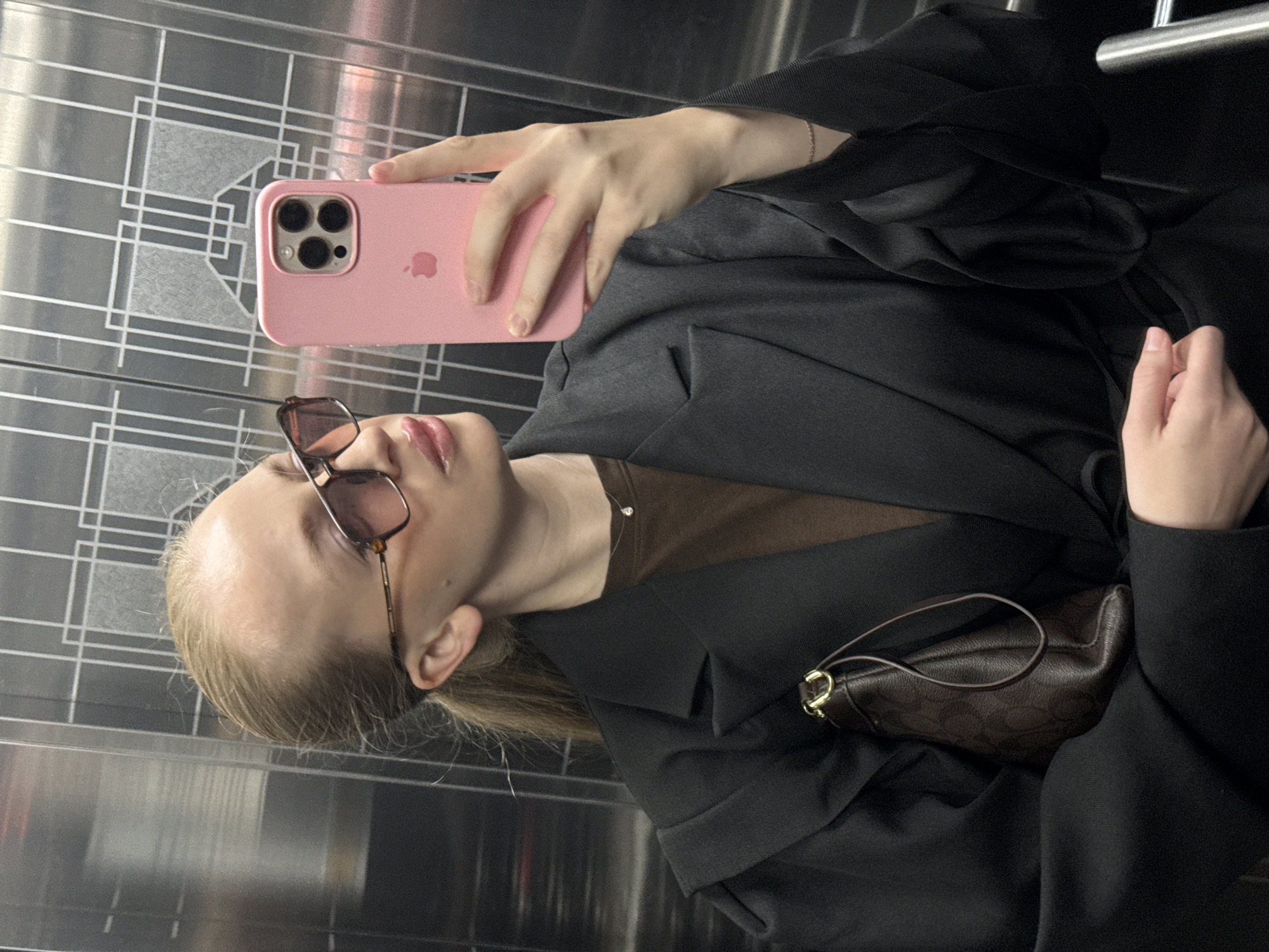 A young woman with blonde hair tied in a ponytail, wearing oversized sunglasses and a black blazer over a brown shirt, stands in an elevator, taking a selfie. She is holding a pink phone in her right hand, which is positioned to capture her reflection in the elevator's metallic door. A small brown purse with a monogram pattern is tucked under her left arm. The elevator's walls have a reflective, metallic finish with a geometric pattern. The location is an elevator in Petropavlovsk-Kamchatsky, Russia.