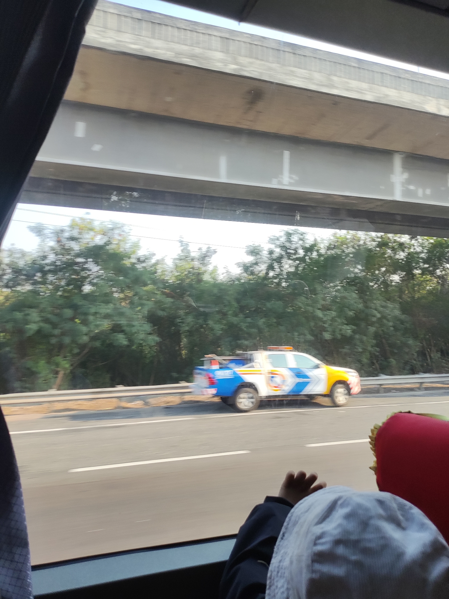 The image shows a side view of a white, blue, and yellow utility truck with a light bar on top, driving on a highway. The truck has a logo on its side that is difficult to discern. The vehicle is driving on the right side of the road. Trees and foliage line the side of the highway, and an elevated highway structure is visible above. In the foreground, from the perspective of someone inside another vehicle, a child's hand and head wearing a white hat are visible, along with a red pillow. The image appears to be taken from a moving vehicle, indicated by motion blur in the foliage and the highway. The scene suggests an outdoor, daytime setting. There is no specific indication of the city of Karawang, Indonesia in the image itself.