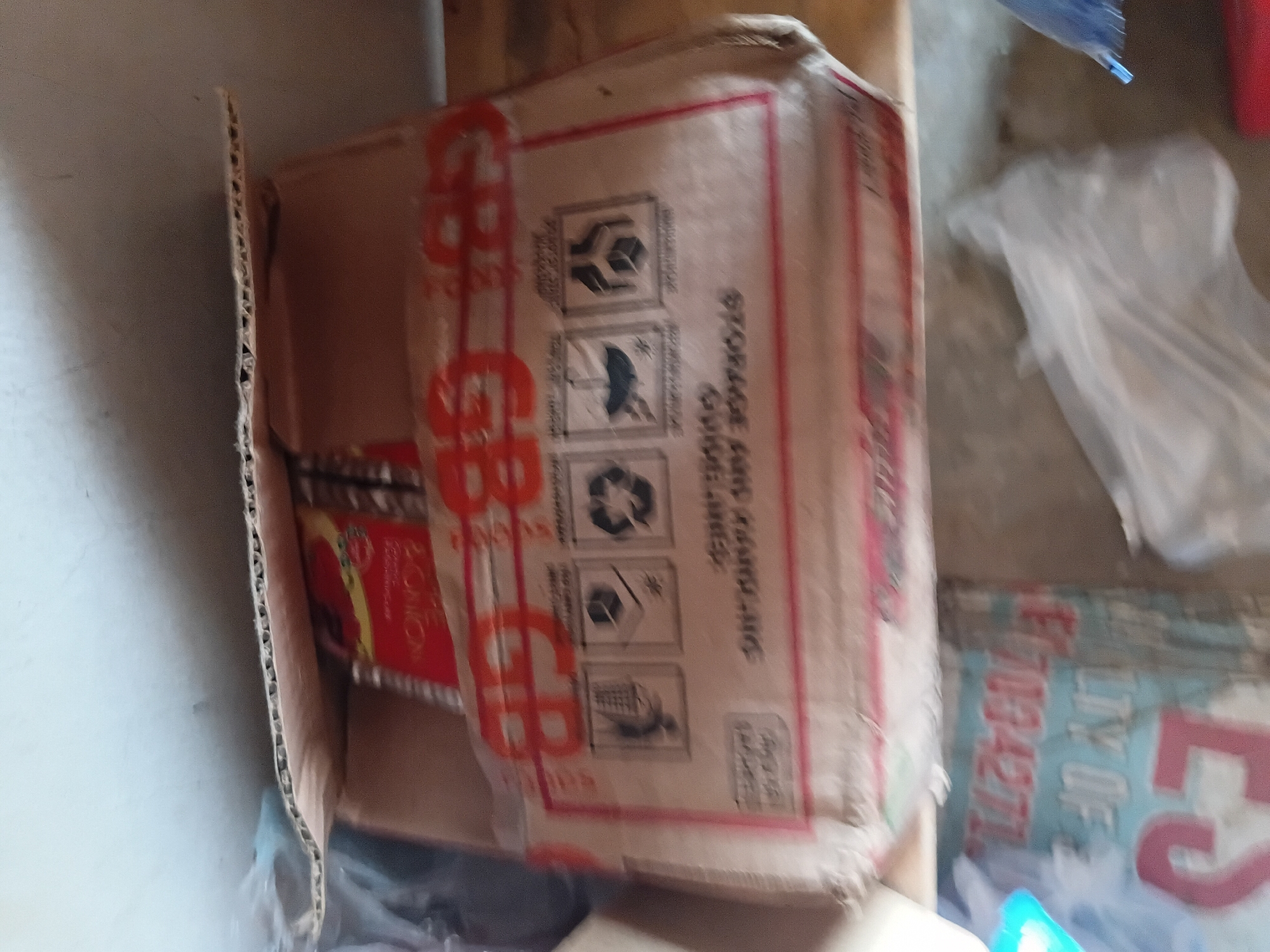 The image shows a close-up, slightly blurry view of a cardboard box with red markings and some text. The box appears to be on its side and is partially open, revealing some contents within.
On the side of the box, there are several printed icons. One icon looks like a recycling symbol, another shows a snowflake or ice crystal, and a third depicts what appears to be a mosquito. Above these icons, text reads "STORAGE AND HANDLING".
The prominent red markings on the box are large letters that appear to spell out "GBB" or similar, repeated. There is also some text in red and yellow on a label visible inside the box, with "SOON" or "BONO" potentially being part of it. On the right side of the image, a blue and red sign is partially visible with the text "TY OF" and a number "80094271" also in red.
The setting appears to be indoors, possibly a storage area or a room. The lighting is artificial and somewhat dim, contributing to the blurriness of the image. There are no people visible in the frame. The overall impression is that of items being stored or packed. The location context of Jalingo, Nigeria is noted but not visually evident from the image itself.