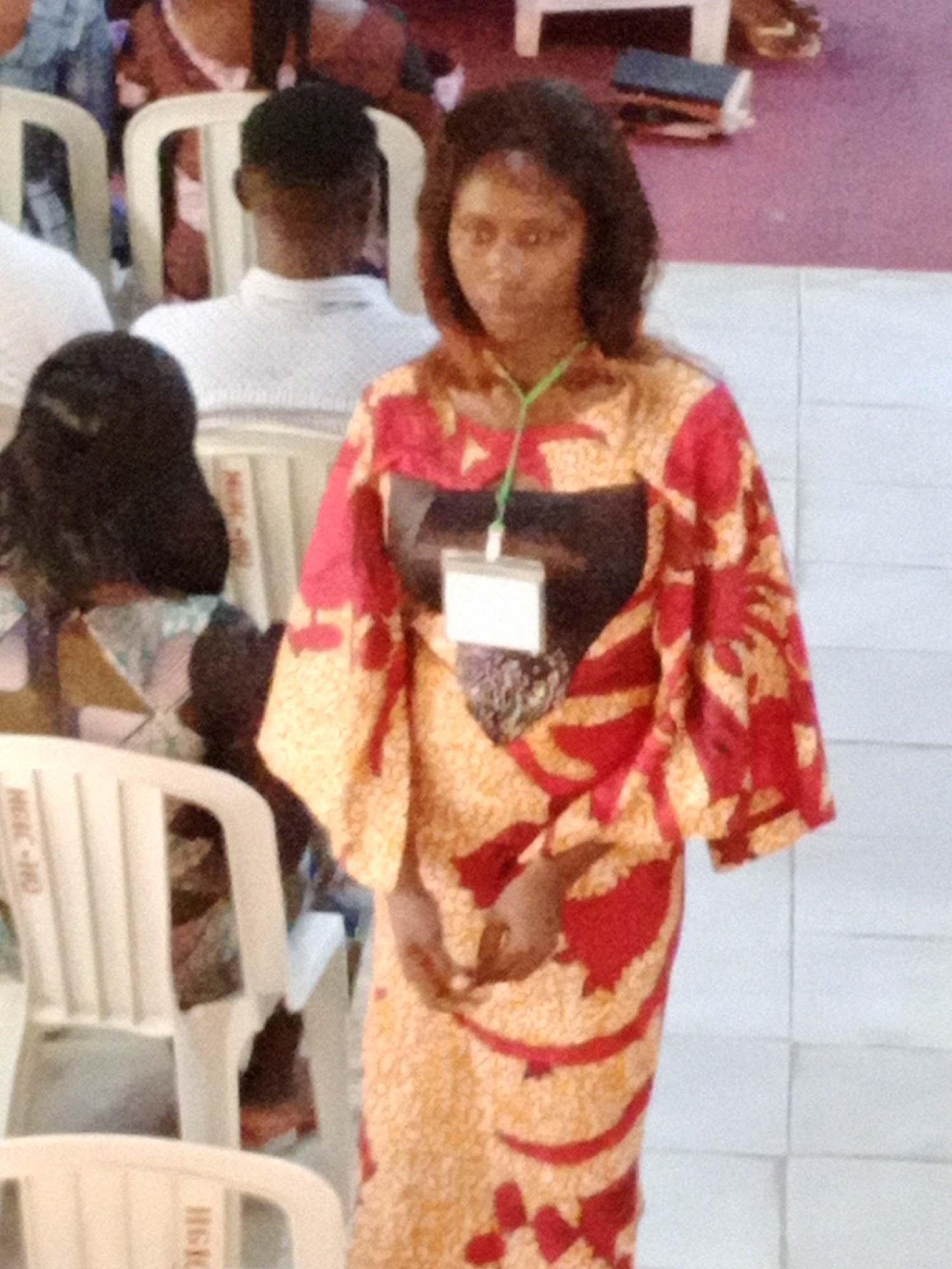 A woman wearing a red and orange patterned dress stands in what appears to be an indoor setting. She has dark hair and is looking towards the camera. A light-colored identification badge hangs around her neck. In the background, several people are seated in white plastic chairs. The floor is tiled with light-colored squares. To the left of the woman, a table with books is visible. The overall scene suggests a gathering or event.
