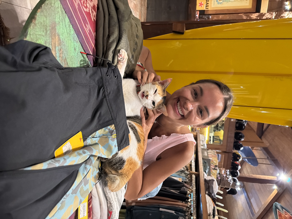 The image displays an indoor scene within a retail establishment, specifically an apparel or surf shop, located in Pecatu, Indonesia. An adult female subject, exhibiting a broad smile and looking directly at the camera, is positioned behind a domestic calico cat. The woman, with dark hair pulled back and wearing a light pink sleeveless top, has both hands gently placed near the cat's head.
The cat, which features white, orange, and black fur patches and a red collar, is lying on a stack of folded clothing atop a low, circular wooden table. Its mouth is open with the tongue slightly visible, and its eyes are squinted, suggesting a vocalization, sneeze, or momentary expression.
In the immediate foreground, a pair of black board shorts with a distinct blue, tan, and grey camouflage-like side pattern, and a yellow retail tag, is prominently displayed on the table. To the left of the cat, a stack of folded dark green fabric items is visible, and to its right, a pile of lighter-colored clothing. The table itself has a distressed, multi-colored painted surface.
The background on the left features a large, bright yellow surfboard propped vertically against a wooden structure, adjacent to a wall-mounted framed item, possibly a map, with "BALI" text visible. On the right side of the background, racks of clothing are discernible, along with an upper shelf displaying multiple dark-colored helmets and a smaller blue surfboard. The ceiling structure reveals exposed wooden beams and artificial lighting fixtures.