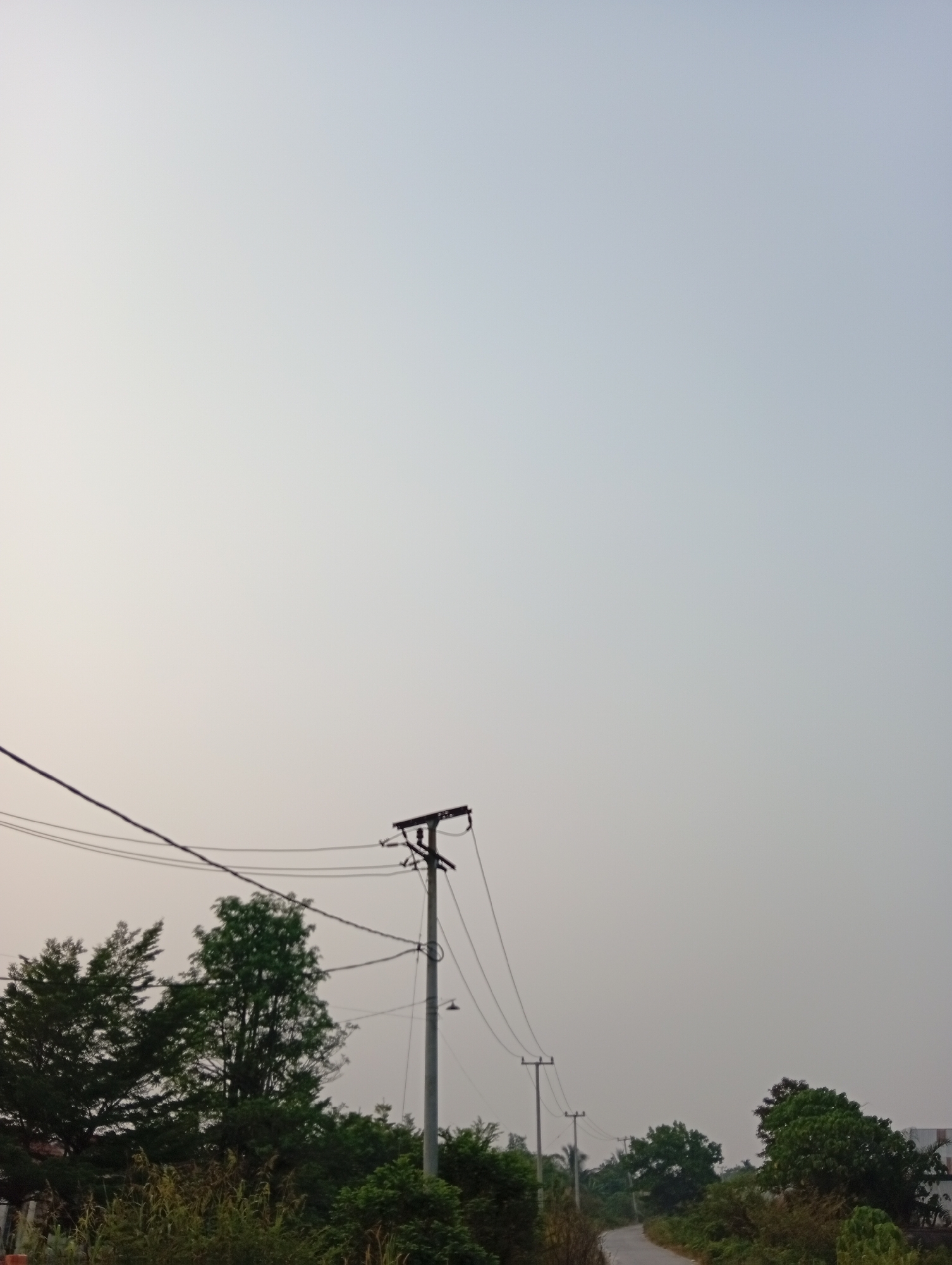 An image captured in Kualu, Indonesia, shows a pale, hazy sky that dominates the upper portion of the frame. In the mid-ground, a gray utility pole stands prominently, supporting multiple electrical wires that stretch across the scene. To the left of the pole, lush green trees are visible, with denser foliage in the foreground. Further to the right, a winding asphalt road recedes into the distance, bordered by vegetation. A second, smaller utility pole is situated further down the road, also with electrical wires. The overall atmosphere suggests a quiet, rural or semi-rural setting with a mild climate.