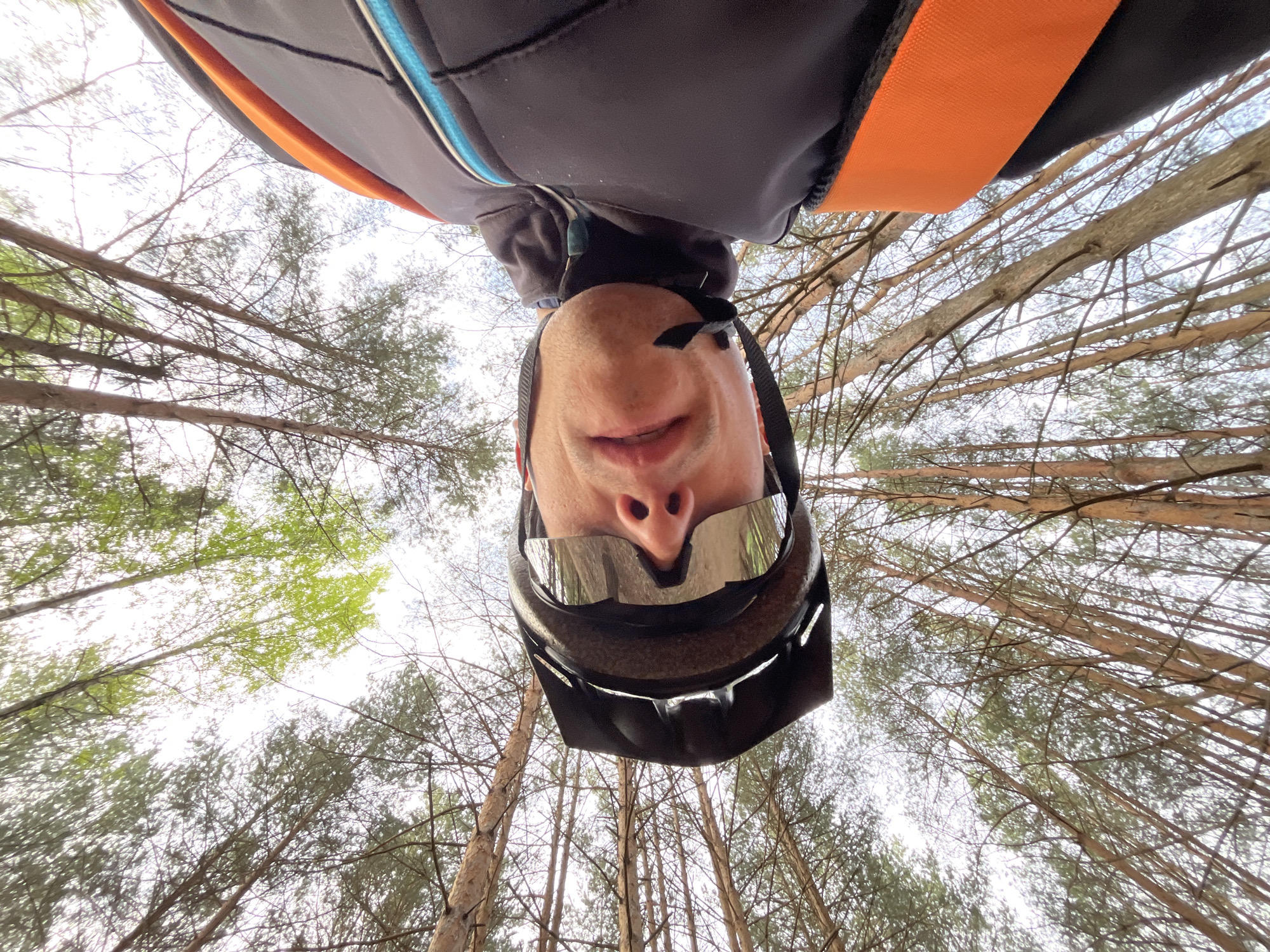The image shows a person wearing a helmet and glasses, looking up from a low angle. The person appears to be in a forest or wooded area, with tall trees and branches visible in the background. The person is wearing a dark-colored jacket with orange and blue accents. The location is likely the city of Chaikovsky, Russia.