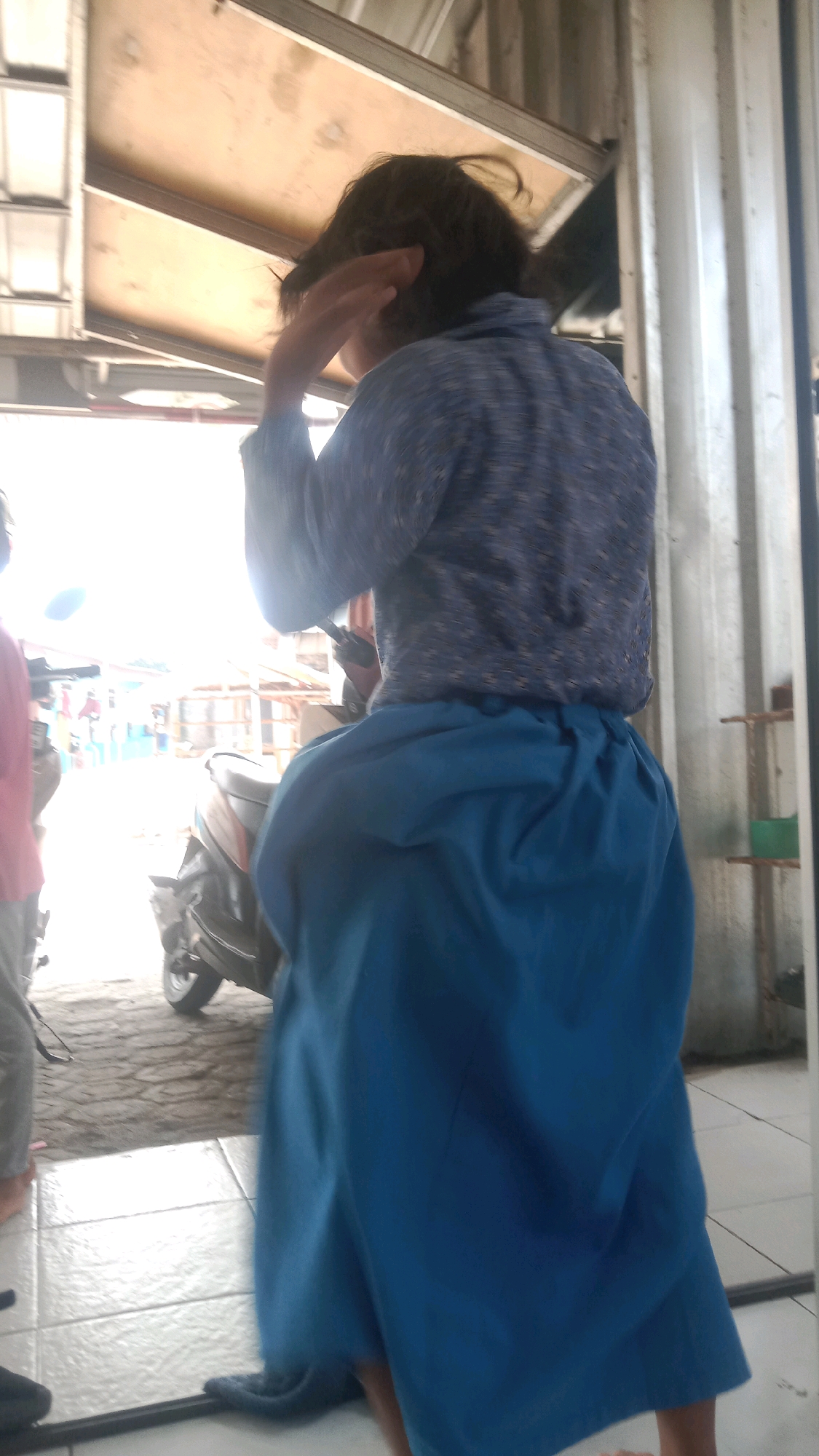 The image shows a person from behind, wearing a blue patterned shirt and a large blue skirt, standing in what appears to be an outdoor market or roadside stall area in Sumberjaya, Indonesia. The person is holding their right hand to their head, possibly adjusting their hair or shielding their eyes from the sun. In the background, a motorcycle is partially visible, and beyond that, there are stalls or buildings with wooden structures and corrugated metal roofing. The ground is a mix of rough pavement and tiled flooring. The overall lighting suggests it is daytime, with bright sunlight casting shadows.