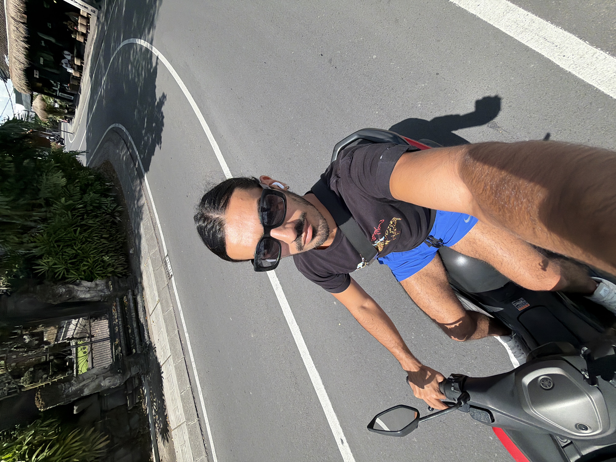 This media file captures a selfie of a man riding a scooter on a sunny day in what appears to be a tropical location. The man is positioned in the foreground, looking directly at the camera with a casual demeanor. He has dark hair styled back, a beard, and is wearing dark sunglasses, a black t-shirt with a graphic design (including an animal, possibly a tiger, and some indistinct text), and bright blue athletic shorts. A black strap, likely from a bag, crosses his shoulder.
He is seated on a grey and black scooter, holding the handlebars with his left hand. A small Yamaha logo is visible on the scooter's front panel. The road is asphalt and curves to the left behind him, marked with a prominent white line along its edge. Clear, sharp shadows from the man, trees, and other objects are cast onto the road, indicating bright daylight.
To the left of the road, there's a sidewalk and a stone wall or fence, beyond which lush green vegetation and palm-like plants are abundant. Further in the background, a building with a traditional thatched roof is visible, enhancing the tropical aesthetic. The overall scene suggests a leisurely ride through a scenic area, consistent with the provided location context of North Kuta, Indonesia. No other specific text is clearly legible in the image.