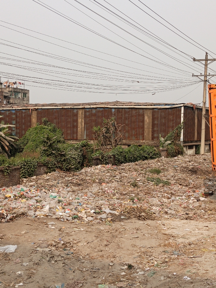 This image depicts a wide-angle view of a heavily littered urban landscape in Boliarpur, Bangladesh, under an overcast sky. The foreground is dominated by an extensive, irregular mound of mixed waste, primarily consisting of plastic bags, bottles, and assorted refuse, covering a dirt ground with scattered debris and dry organic matter. No individuals are present.
In the midground, a low, weathered brick wall runs horizontally, topped by dense, unruly green vegetation, including bushes and vines. Behind this foliage stands a long, single-story building constructed from vertically oriented, heavily rusted corrugated metal sheets. Its flat roof, also appearing to be made of corrugated material, shows signs of weathering.
The background features a multi-story, light-colored residential or commercial building on the left, with laundry visible hanging on its rooftop. Numerous black utility lines crisscross the upper portion of the frame against a uniform light grey sky, originating from a utility pole partially visible on the far right. An orange shipping container is positioned vertically on the far right edge of the frame, adjacent to the utility pole.
