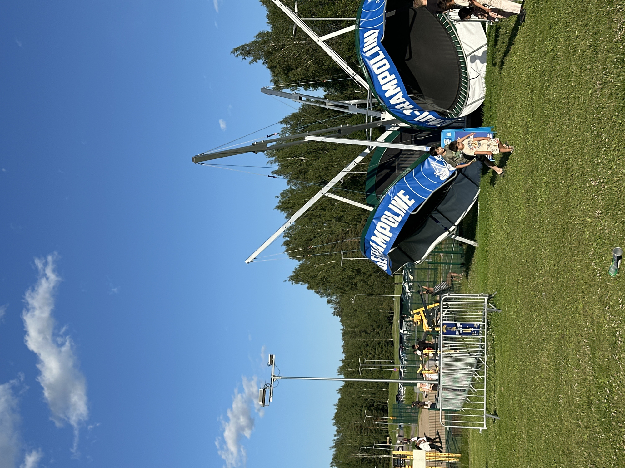 This media file depicts an outdoor scene on a sunny day, with a clear blue sky and scattered white clouds. The primary focus is a bungee trampoline attraction. Two large bungee trampolines are visible, one partially obscured by the other. Both feature a black jumping surface and a green safety net. Blue banners with white lettering reading "JUMPLINE" are prominently displayed on the structures.
Several people are present, interacting with the attraction and its surroundings. In the foreground, two young boys are standing near a metal barricade that appears to be part of the queue system for the bungee trampoline. One boy is wearing a green t-shirt and the other a light-colored t-shirt with a graphic design and patterned shorts. Further in the background, other individuals can be seen near the trampolines, some appearing to be waiting or observing.
The setting is a grassy area, likely a park or an outdoor recreational facility. Trees line the background, suggesting a natural or semi-natural environment. Several metal poles with lights are also visible, possibly for illumination or security. The overall atmosphere appears casual and recreational, suggesting a day of fun and outdoor activity. The bright sunlight indicates it is likely daytime.