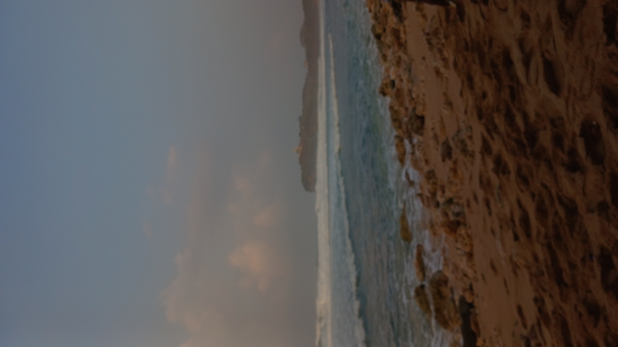The image depicts a beach scene, likely in Tepus, Indonesia, under a hazy sky. The foreground is dominated by a sandy beach with scattered rocks. In the midground, the ocean features white-capped waves rolling towards the shore. A distant headland, possibly with structures on it, is visible on the horizon. The overall atmosphere is subdued, suggesting it might be late afternoon or early morning. There are no people or specific activities clearly discernible in the image. The lighting is soft and diffused, contributing to the hazy, muted tones of the scene.