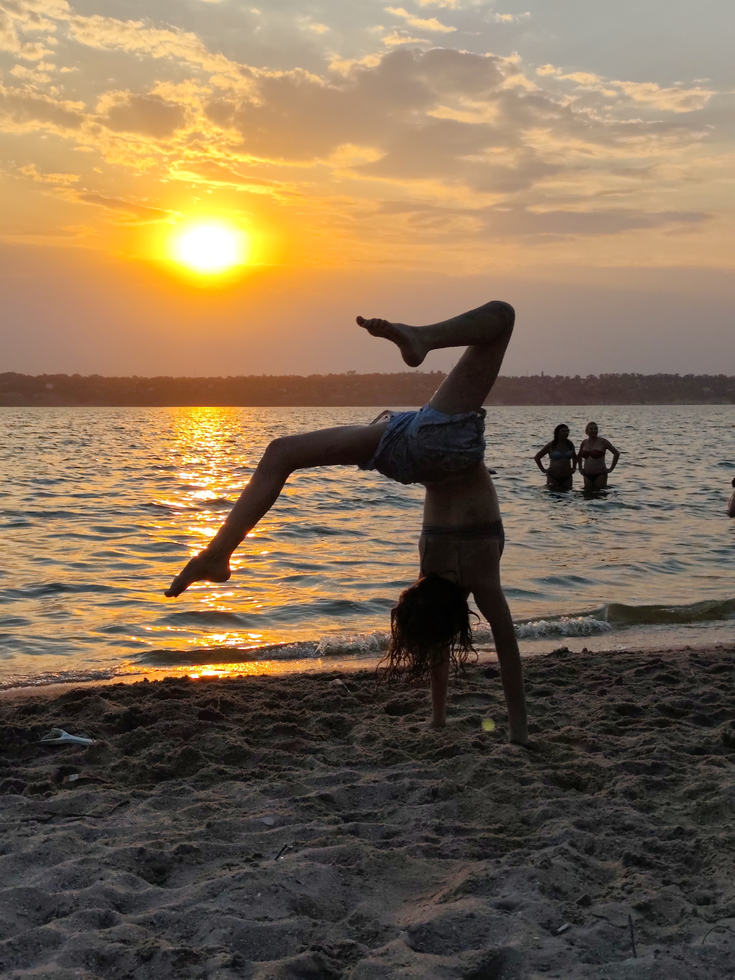 At sunset, on a sandy beach, a young person is performing a handstand in the shallow water's edge, with their legs extended upwards and bent at the knees. The sun is low on the horizon, casting a warm orange glow across the sky and reflecting brightly on the rippling water. In the background, two women stand in the water, partially submerged. The scene is likely a beach on the Black Sea coast near Mykolaiv, Ukraine, given the commonality of such recreational activities during sunset hours in coastal regions.