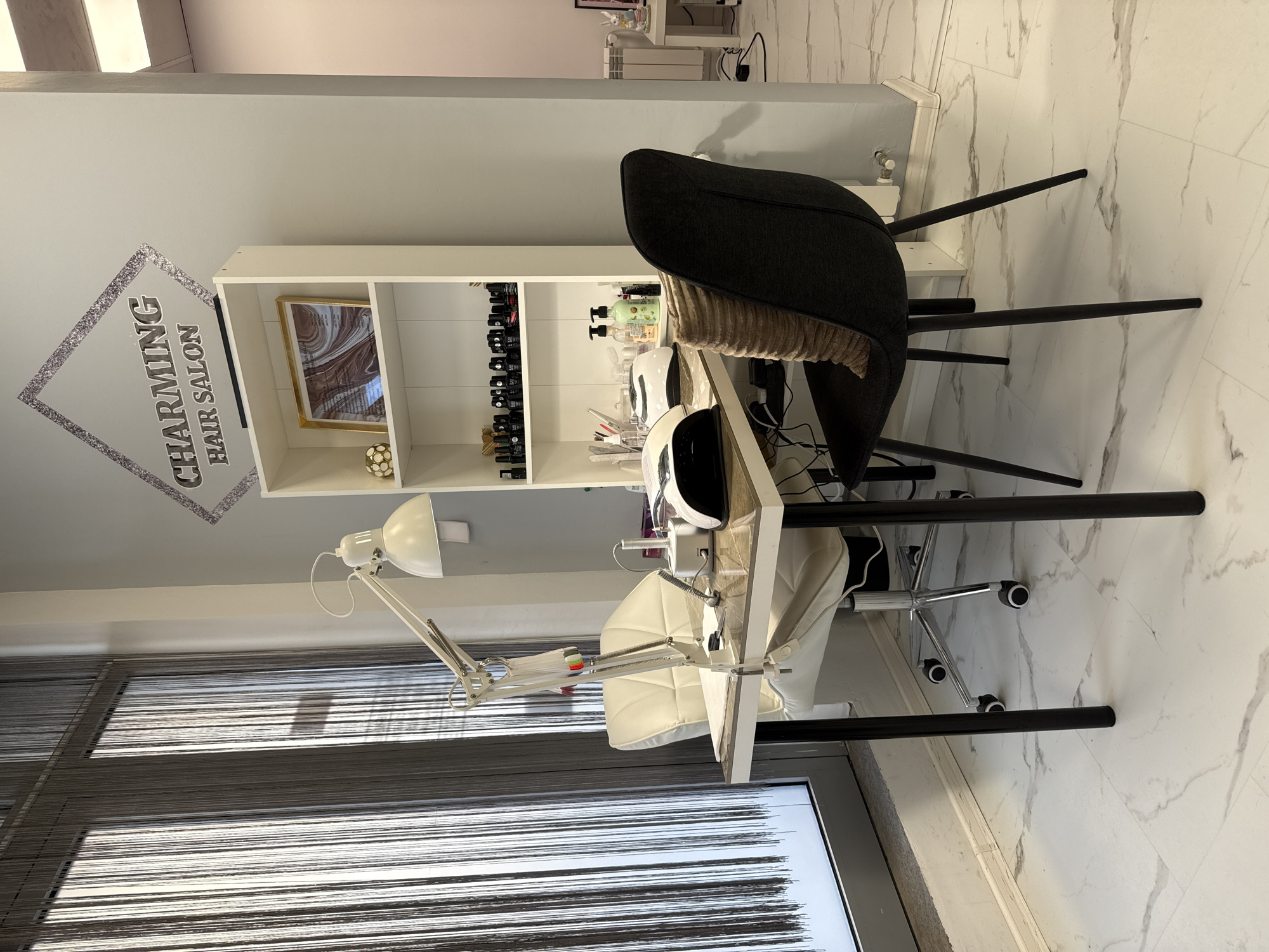The image depicts a hair salon interior. A white bookshelf is against a wall, displaying items such as nail polish bottles, lotions, and framed art. A black chair with a tan cushion is positioned near a manicure table. The table is equipped with a white lamp and a UV nail dryer. A white chair with a wheeled base sits at the table. The scene is illuminated by the lamp and natural light coming through a window with string curtains. The wall features a logo that reads "Charming Hair Salon." The floor is white marble. Location in the city of Sofia, Bulgaria cannot be determined from the image.