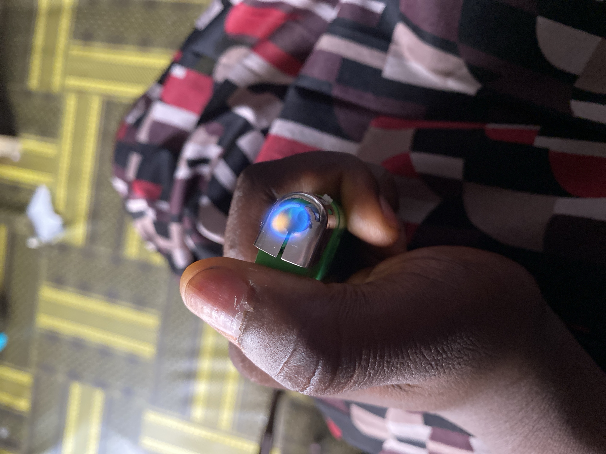 The image shows a close-up of a person's hand holding a green lighter that is lit, with a blue and yellow flame visible. The person's skin appears dark, and their fingernails are visible. The background is blurred, but suggests a patterned fabric, possibly clothing, and a floor with a yellow grid pattern. The overall lighting suggests an indoor setting, possibly at night or in a dimly lit room. There is no visible text in the image. The context of Jalingo, Nigeria is provided but not explicitly visible in the image itself.