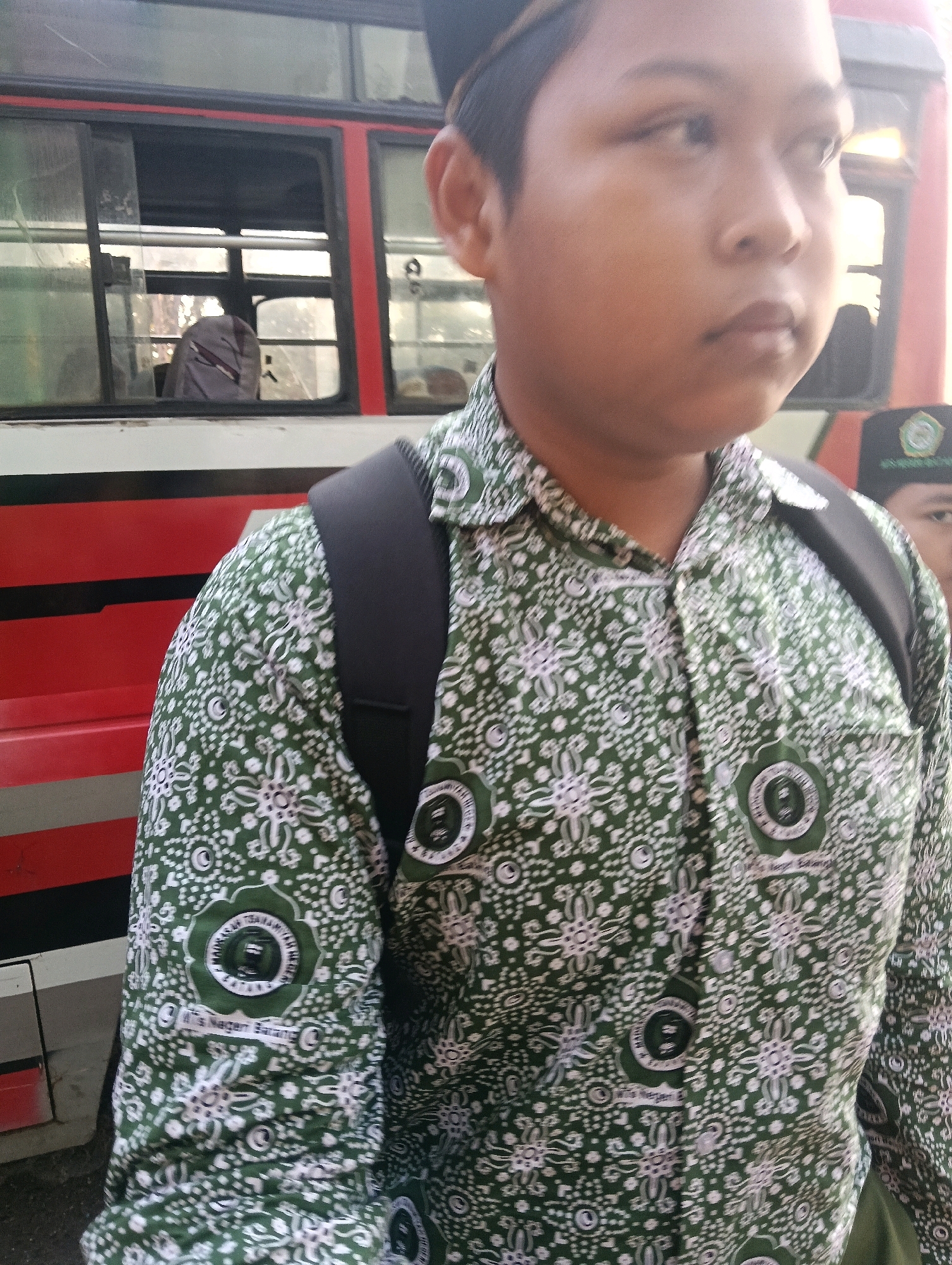 The image depicts a close-up view of a young person wearing a green and white patterned uniform shirt, a black backpack, and a black cap with a logo. The person is standing in front of a red and white bus. The bus has windows, one of which shows a seat. The background is slightly blurred, but appears to be an outdoor environment, possibly a street or a parking area. There are hints of the location "Adinuso, Indonesia" on the person's uniform, specifically on a circular emblem on the left side of the shirt which reads "MTs NEGERI 1 BANJARNEGARA" and below it "Adinuso, Banjarnegara". The lighting suggests it is daytime.