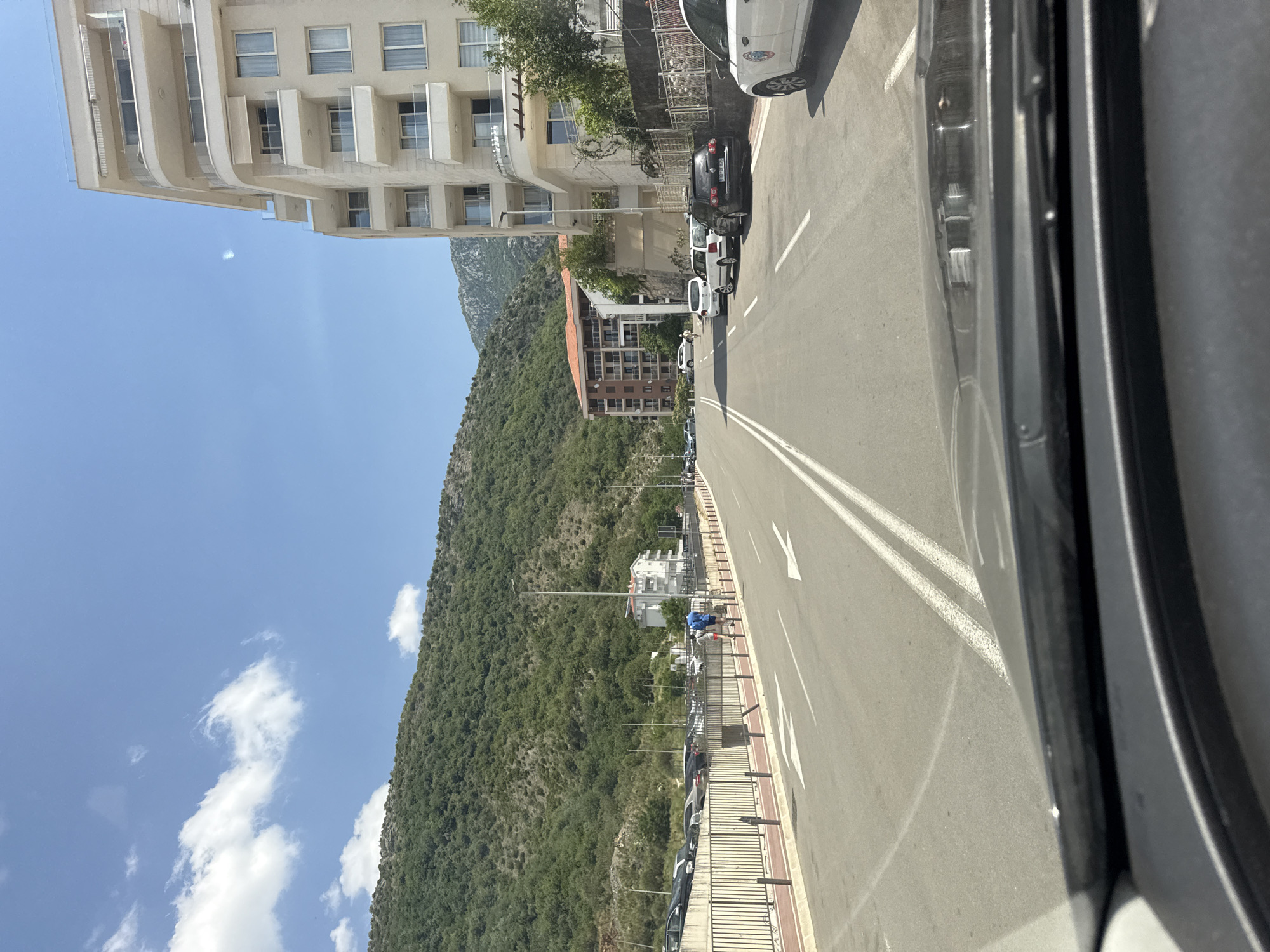 A street scene in Rafailovići, Montenegro, shows a road with double white lines in the center. On the right, a modern, light-colored multi-story building with balconies and awnings lines the street. Parked cars are visible along the right side of the road, including a white car with decals. Further down the street, other vehicles are parked, and a dark SUV is visible. Across the road, a pedestrian walkway is bordered by a metal fence. Beyond the walkway, a parking lot is filled with cars. A steep, tree-covered mountain slopes upwards in the background. A few white clouds dot the bright blue sky. A person in a blue shirt and shorts is walking on the sidewalk.
