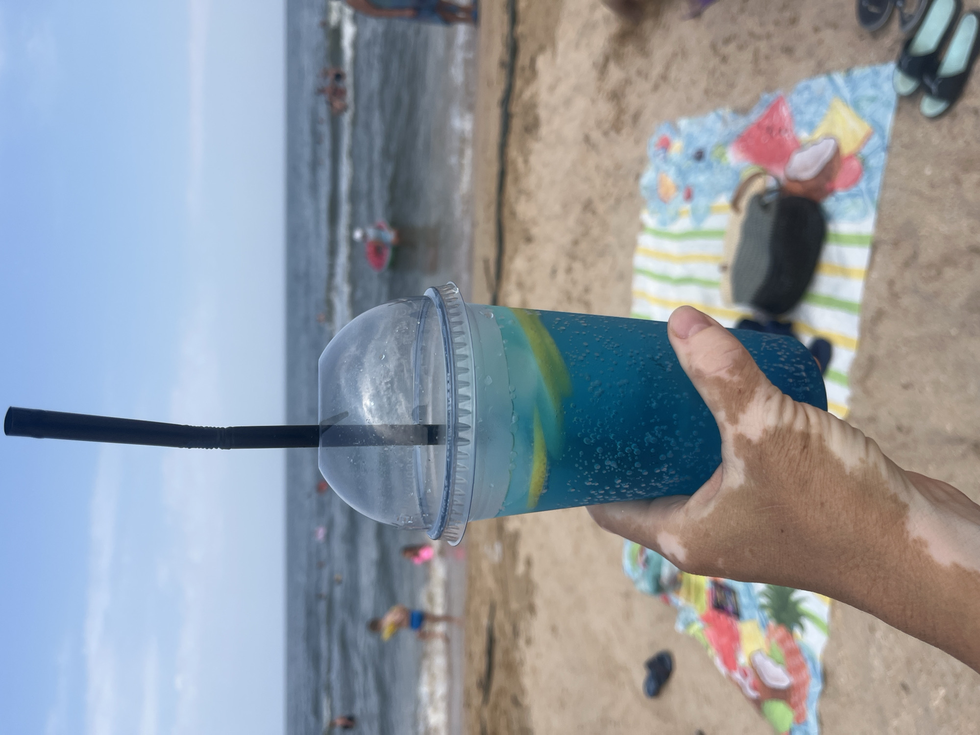 The image shows a person's hand holding a plastic cup of blue liquid with ice and lemon slices, with a black straw protruding from the top. The background is a beach scene with the sea and people swimming or relaxing in the water. A striped towel with colorful patterns is spread on the sand in the foreground, along with a dark bag and a pair of black flip-flops. The hand holding the cup has patches of depigmentation. The location is identified as Golubitskaya, Russia, a Black Sea resort known for its beaches.
