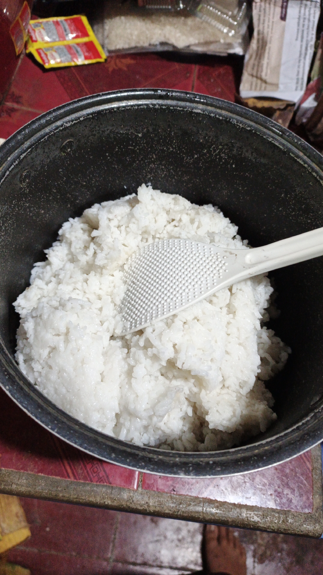 A medium shot, taken from a slightly elevated angle, shows a black rice cooker filled with cooked white rice. A white plastic rice paddle with a textured surface rests on top of the rice. The rice cooker is placed on a red, tiled surface. In the background, out of focus, are packets of what appear to be seasoning or instant noodles, along with bags of uncooked rice. The overall scene suggests a domestic kitchen environment. The location of Gunungreja, Indonesia is provided as context, but is not visually discernible from the image itself.