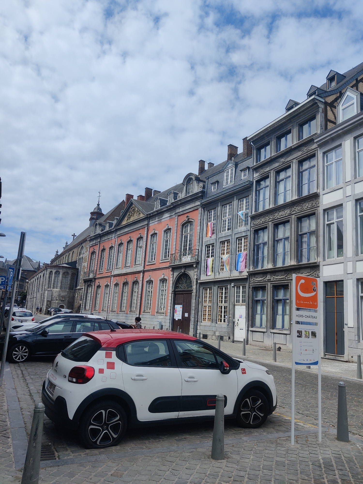 The image captures a street scene in Liège, Belgium, featuring a row of historic buildings with ornate facades and mansard roofs. The architecture suggests a European city with a rich historical past. In the foreground, a white car with a red roof is parked on a cobblestone street. The car has a logo with red squares on its side and a Turkish flag emblem on the front fender. A black car is parked behind it. Further down the street, several other cars are visible, and pedestrians are present. The buildings are adorned with decorative elements, including balconies and numerous windows. Flags, possibly representing different regions or events, are hung from some of the buildings. A signpost with an orange circle logo and text indicating "CAMBIO HORS-CHÂTEAU" is prominently displayed on the right side of the image, suggesting a car-sharing service or a point of interest in the Hors-Château district of Liège. The sky is cloudy, indicating an overcast day.