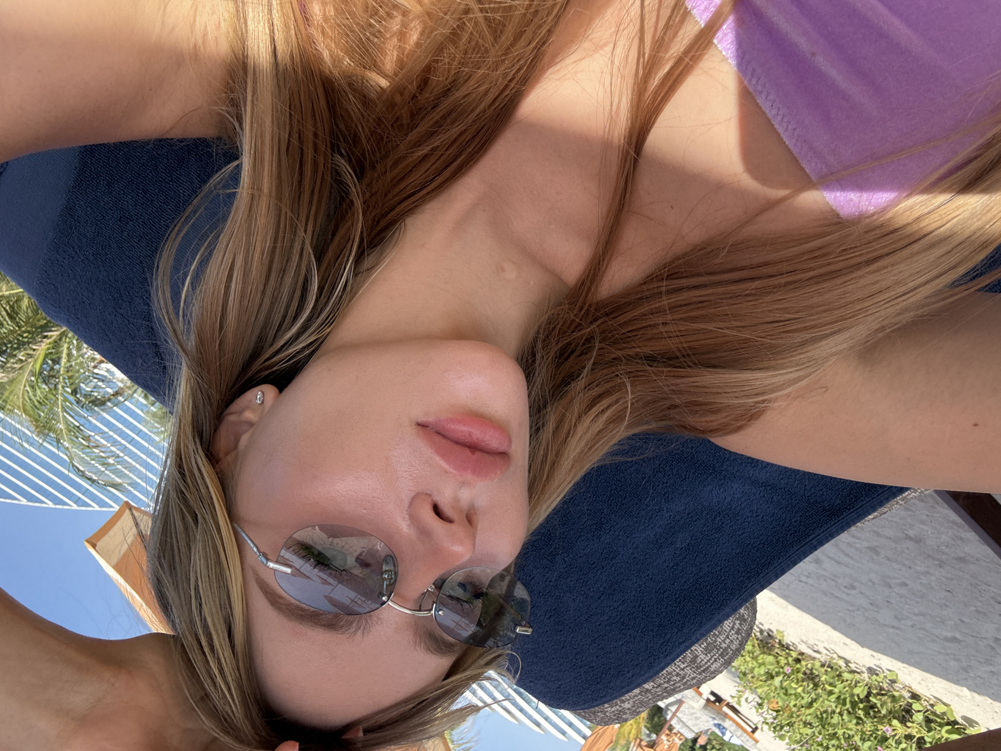 A woman with light brown hair is laying on a navy blue towel, likely on a lounge chair. She is wearing round sunglasses with silver frames. A lavender fabric, possibly a swimsuit, is visible at the top right. The image appears to be taken outdoors on a sunny day. In the background, there is a tall building with a distinctive striped pattern, indicative of modern architecture in Dubai. A palm tree and other greenery are also present. The setting suggests a resort or beachside location in Dubai, United Arab Emirates.